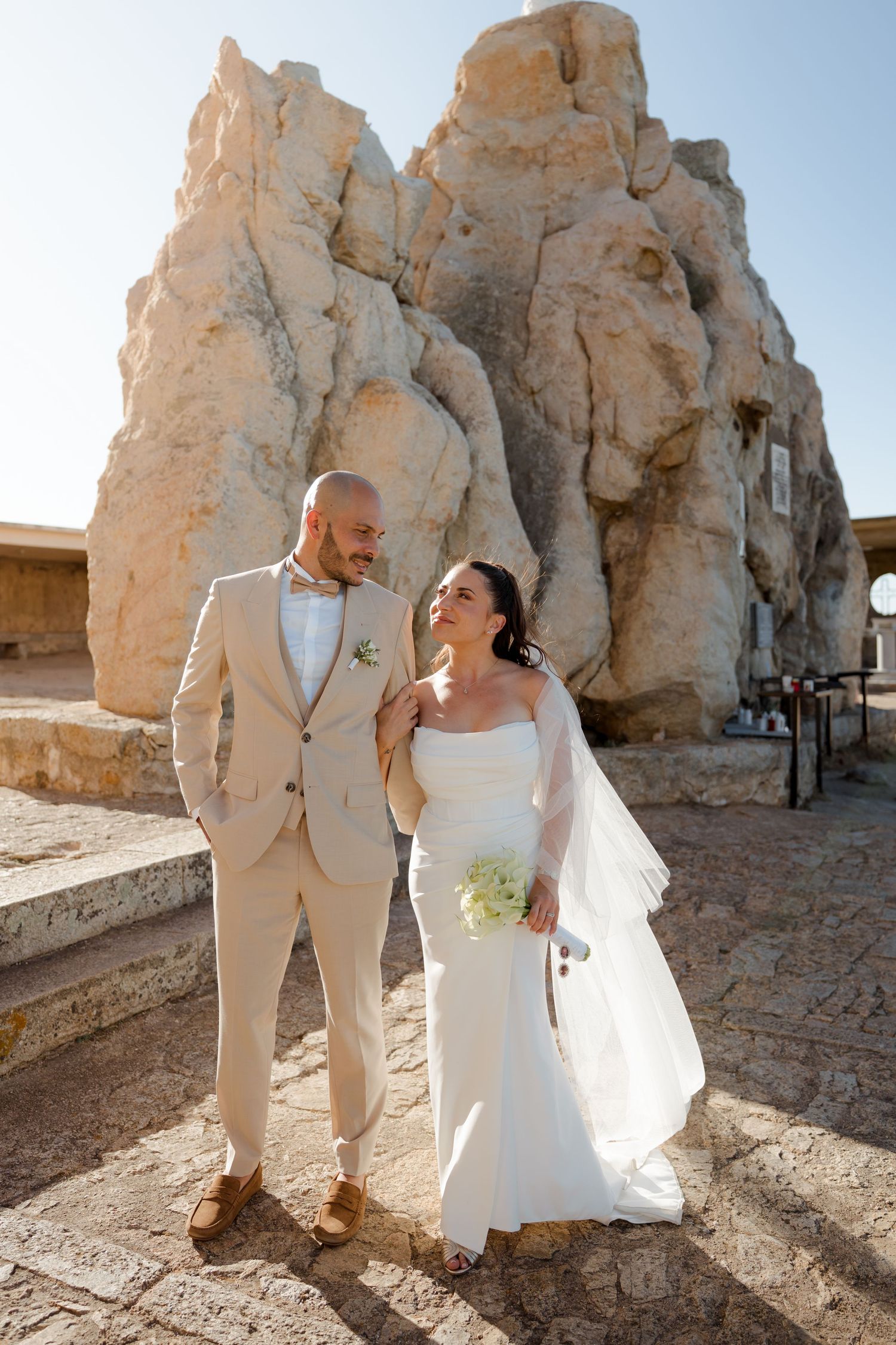 Smiling newlyweds in beige suit and white gown pose together against sunlit rocky formation backdrop.