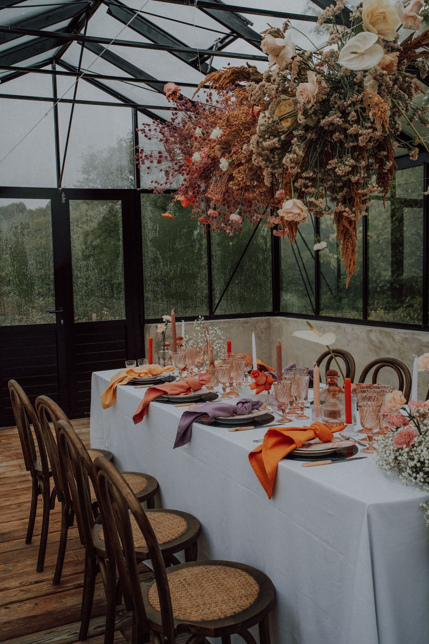 Stunning greenhouse wedding reception table with hanging floral installation and colorful candles and napkins.