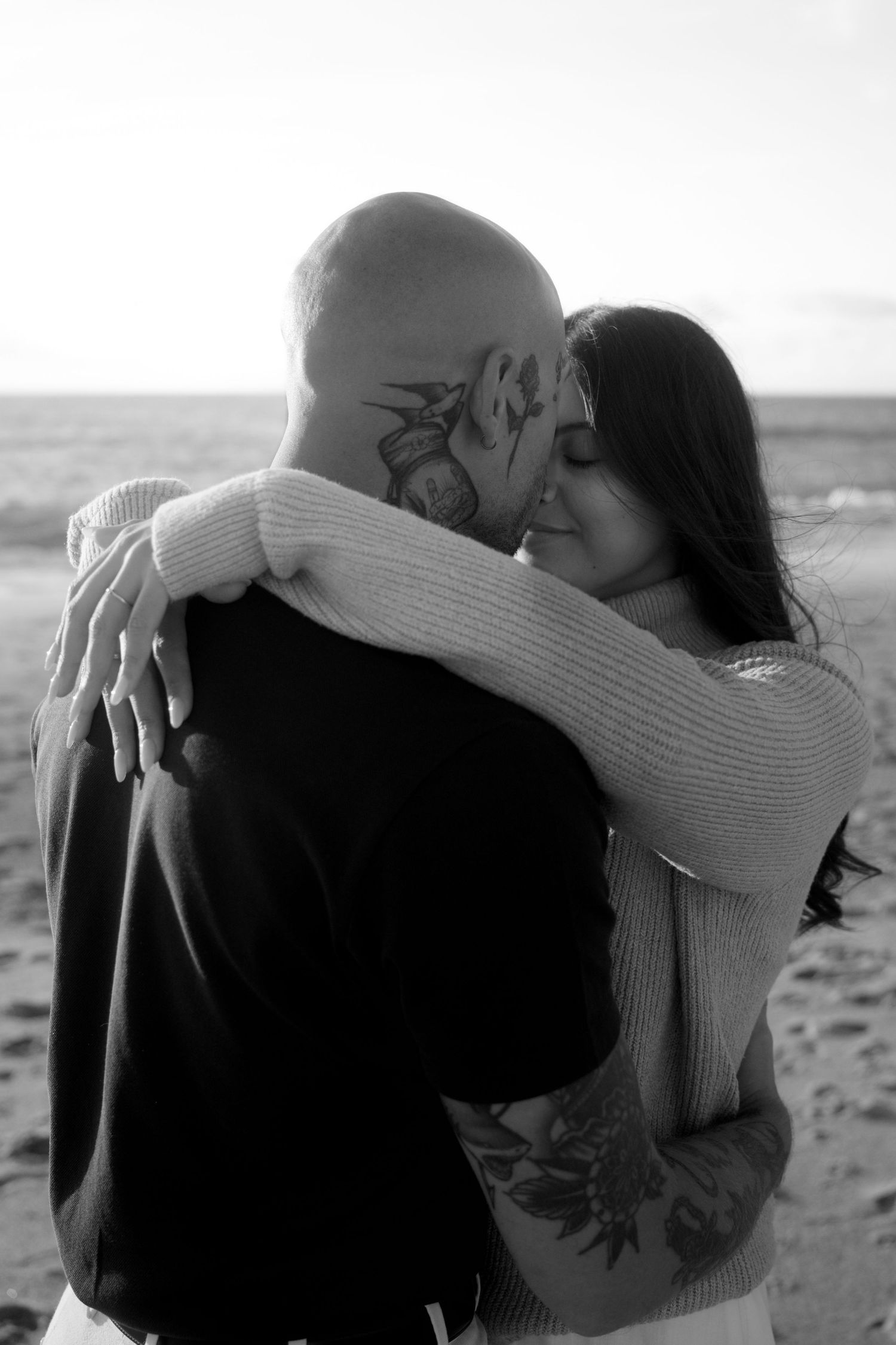 Black and white photo of couple hugging on beach, both with visible tattoos, ocean in background.