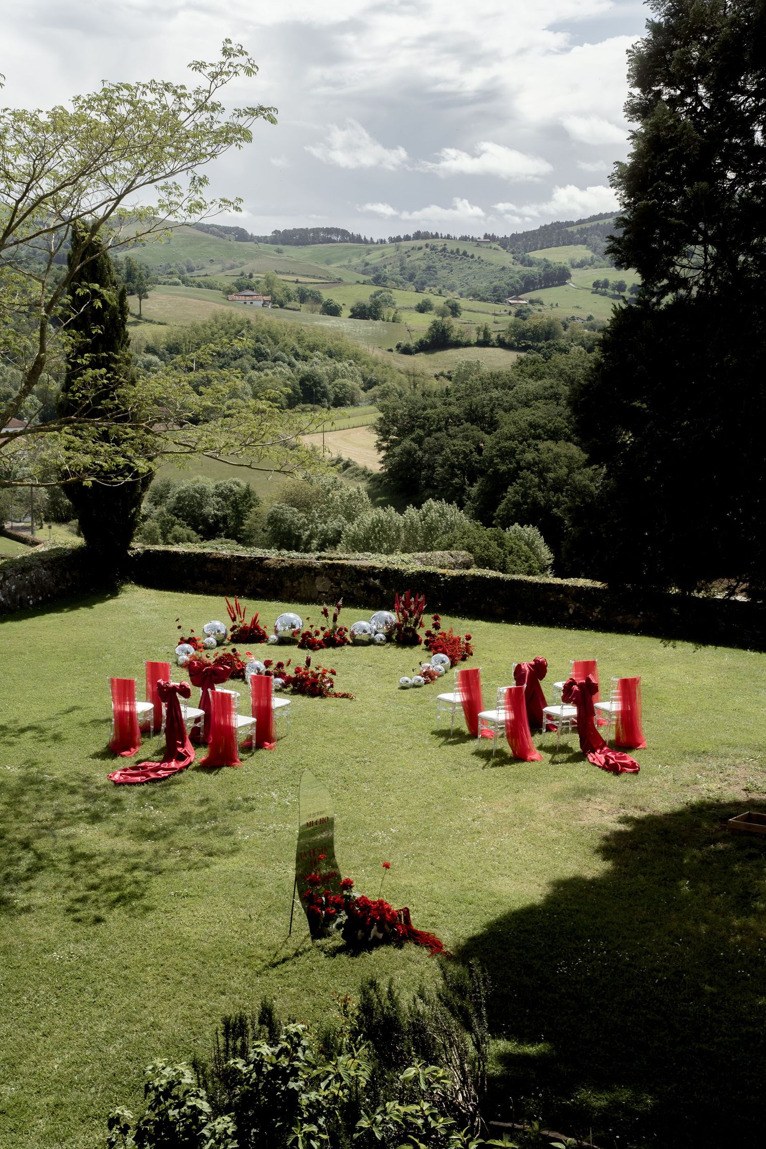 Outdoor wedding ceremony setup with red chairs and floral arrangements on green lawn with rolling hills backdrop.