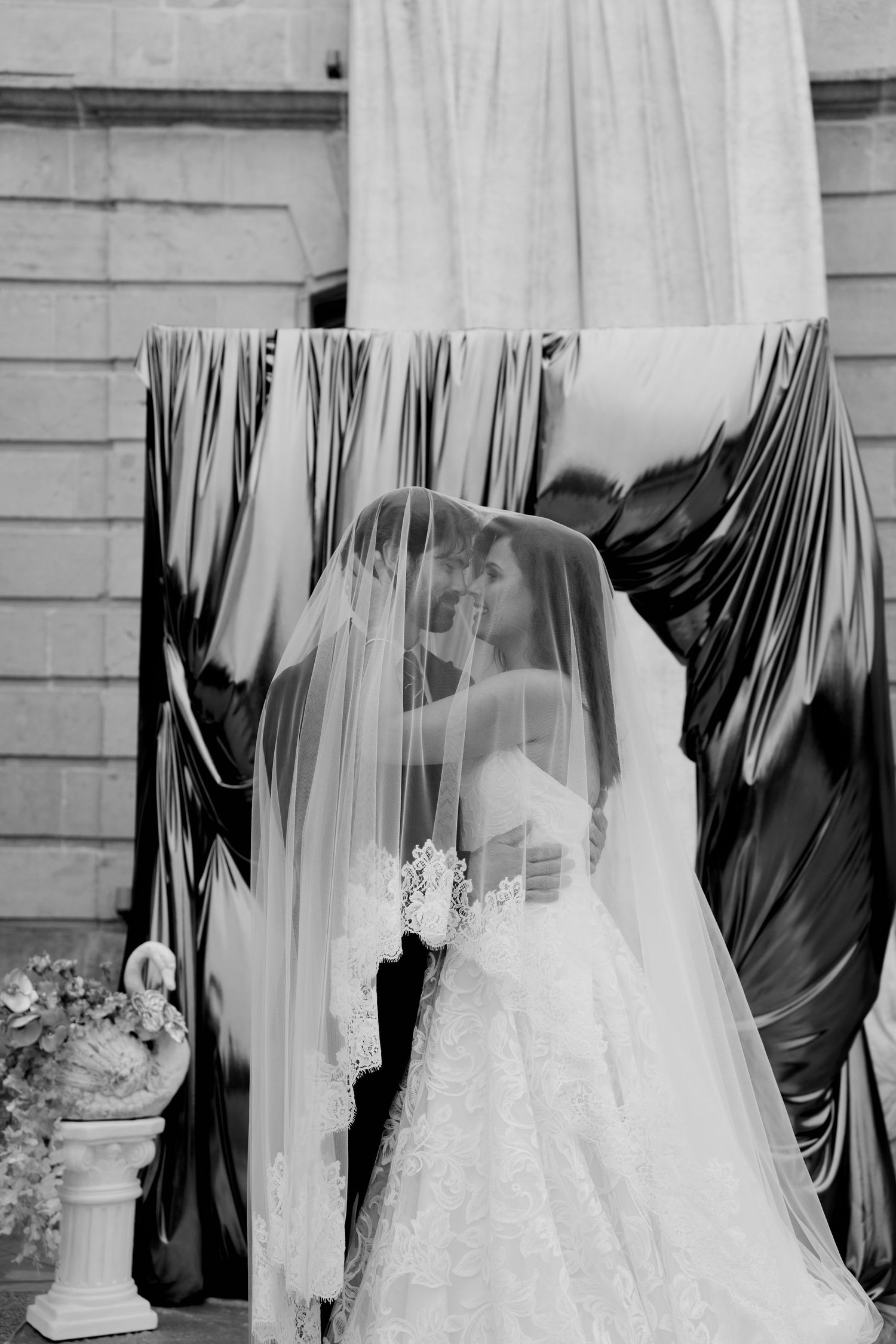 Black and white photo of bride and groom sharing intimate moment under wedding veil at ceremony.