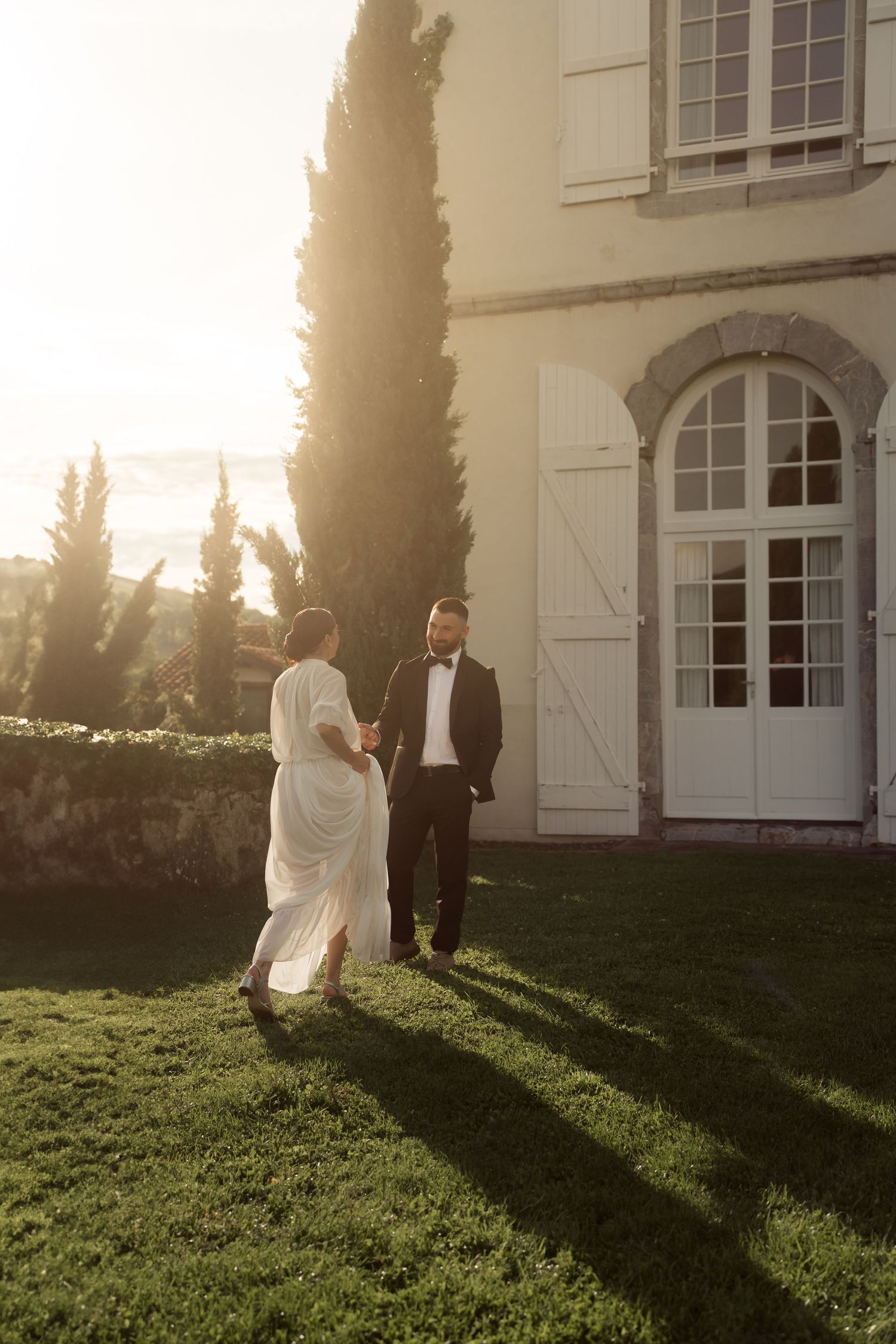Bride in flowing white gown walks toward dapper groom outside elegant French chateau at golden sunset.