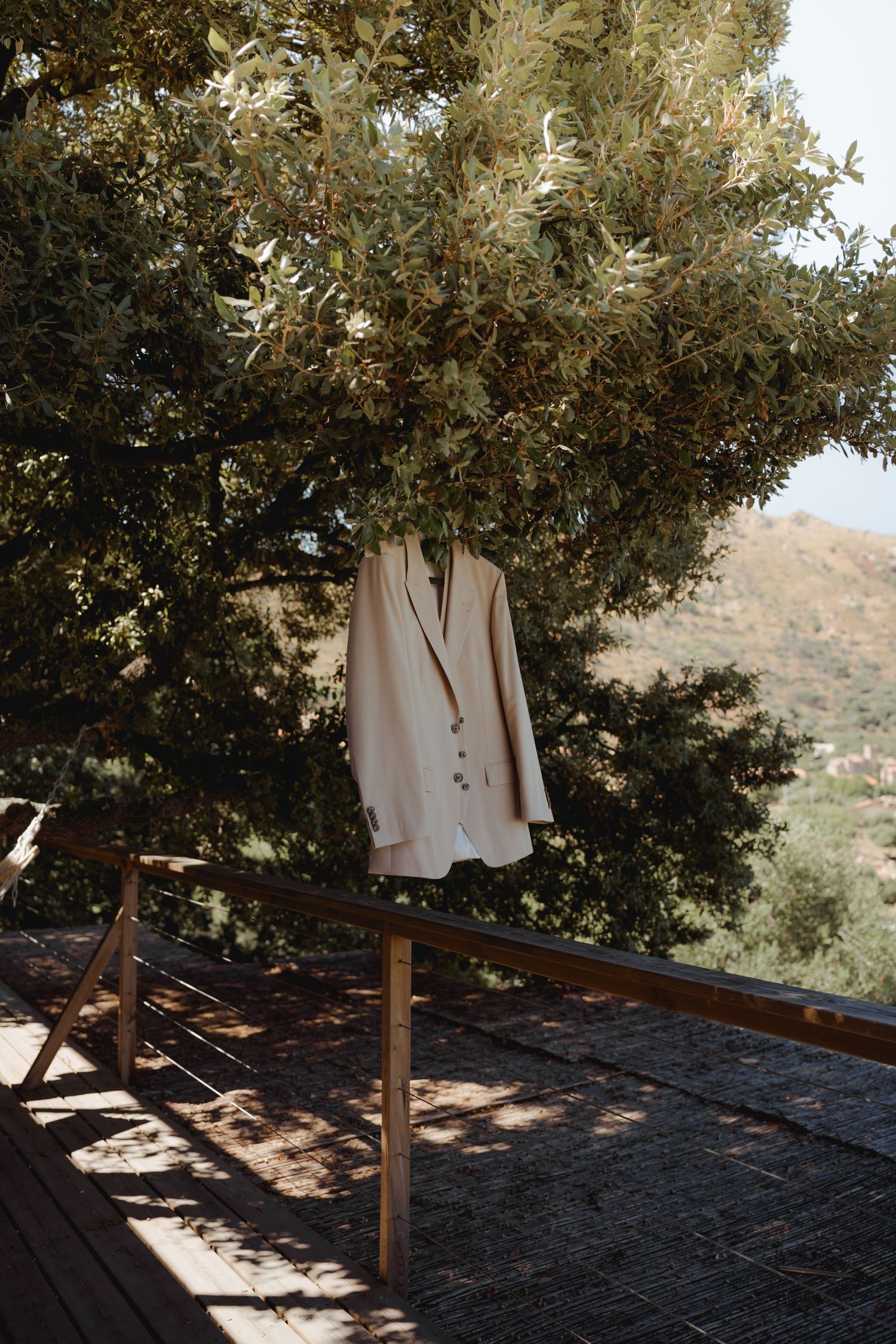 Cream blazer hanging from olive tree branch on wooden deck with mountain views.