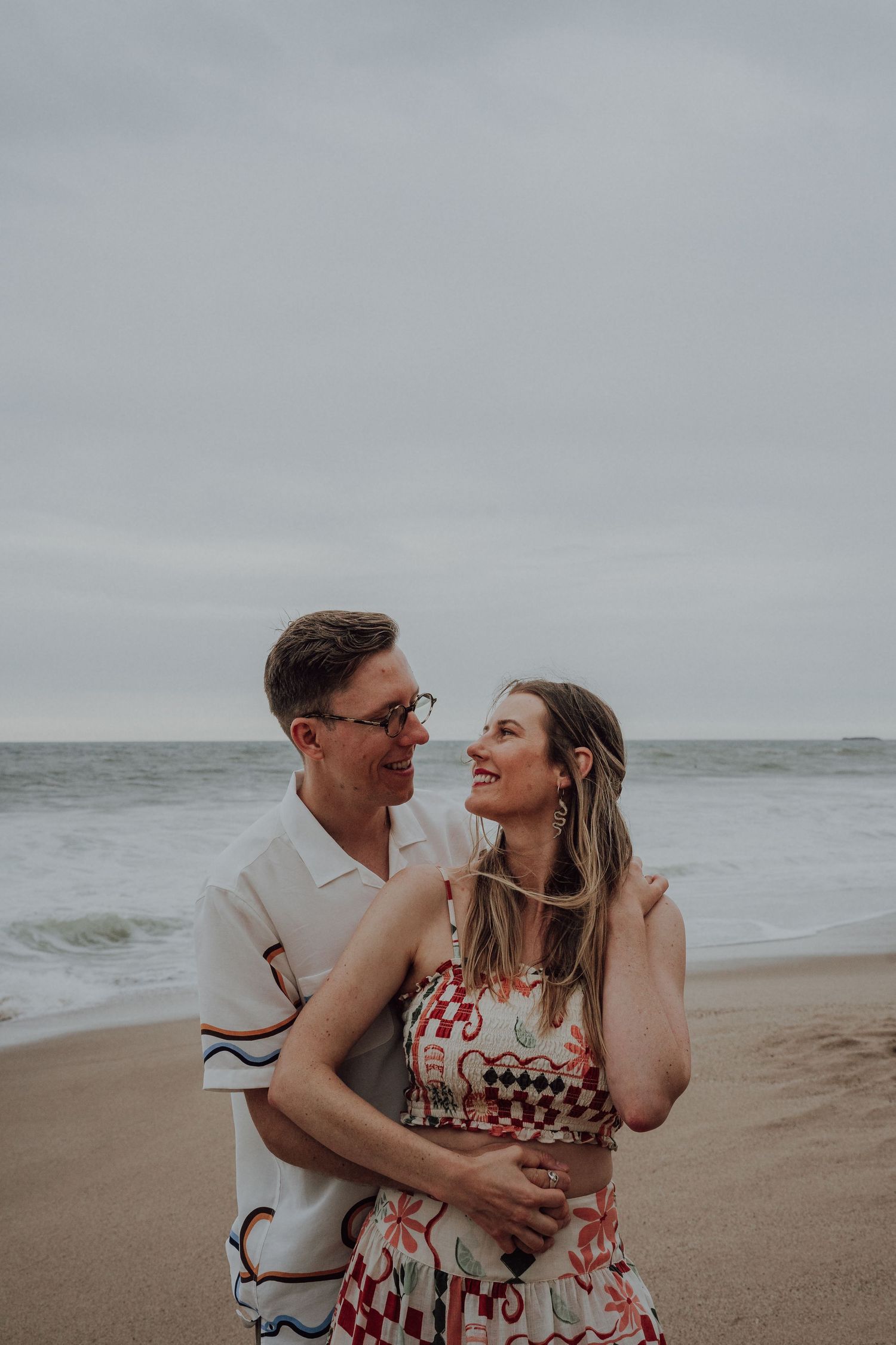 A smiling couple embraces on a sandy beach, the woman in a floral dress and man in a white shirt.