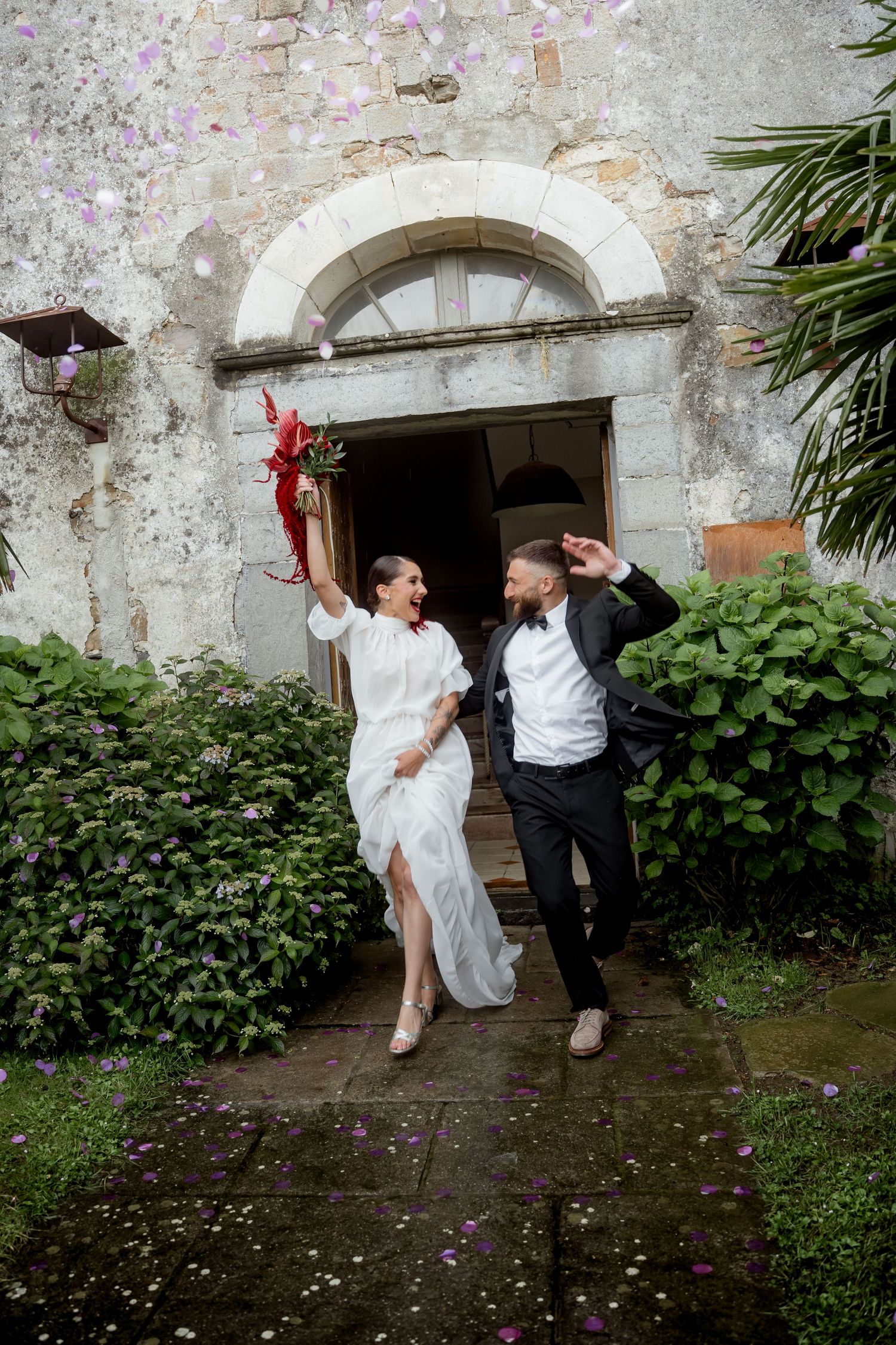 Laughing bride and groom exit rustic stone church with red bouquet amid falling confetti.