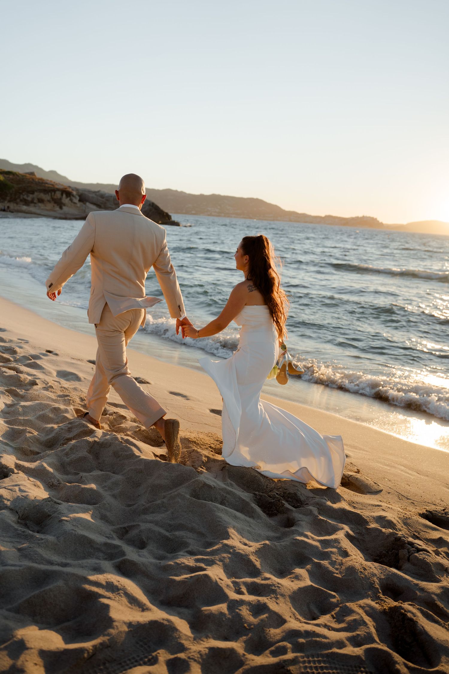 Wedding couple holding hands walking along sandy beach shoreline at golden hour sunset.