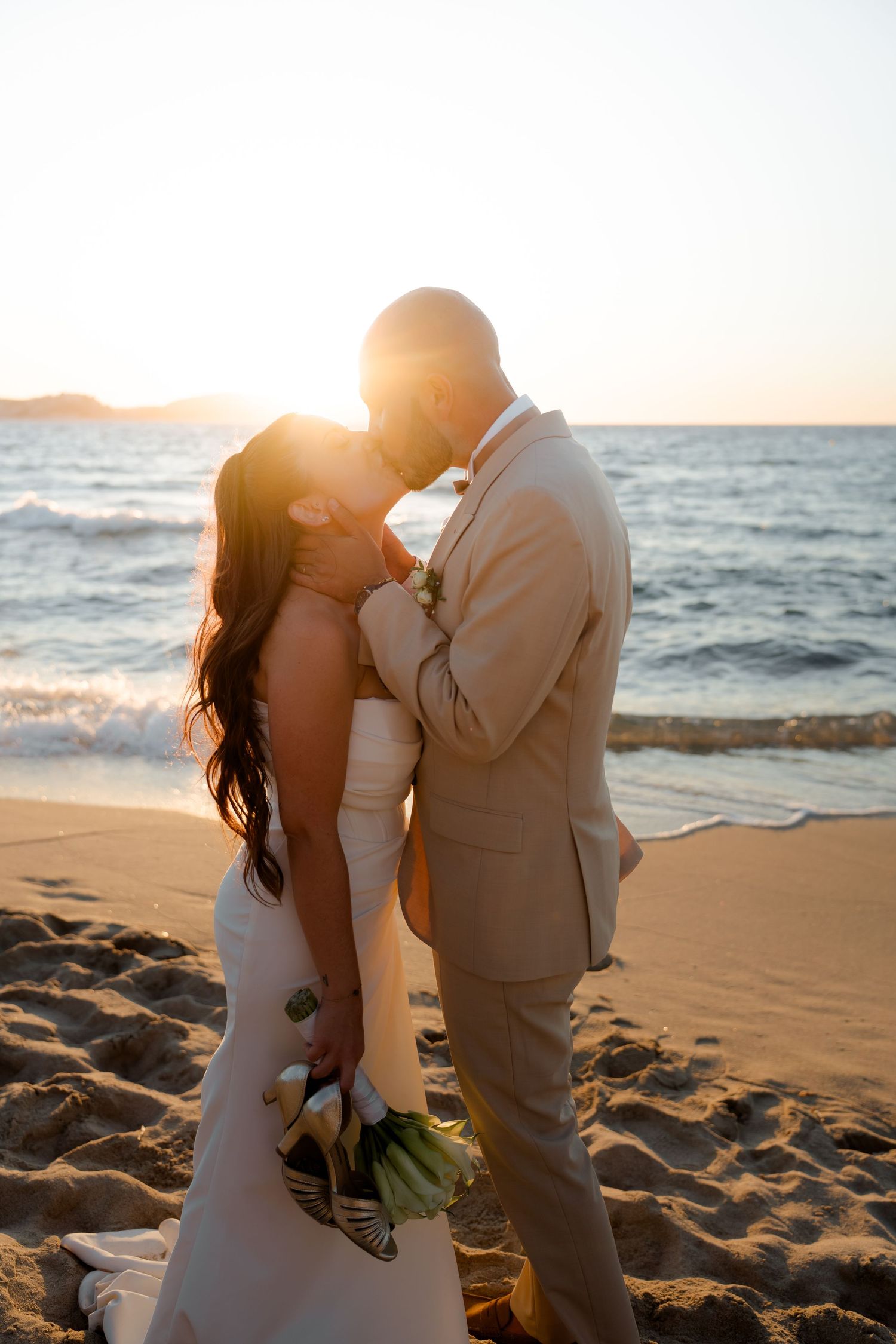 Bride and groom sharing romantic sunset kiss on beach, bride holding white floral bouquet.