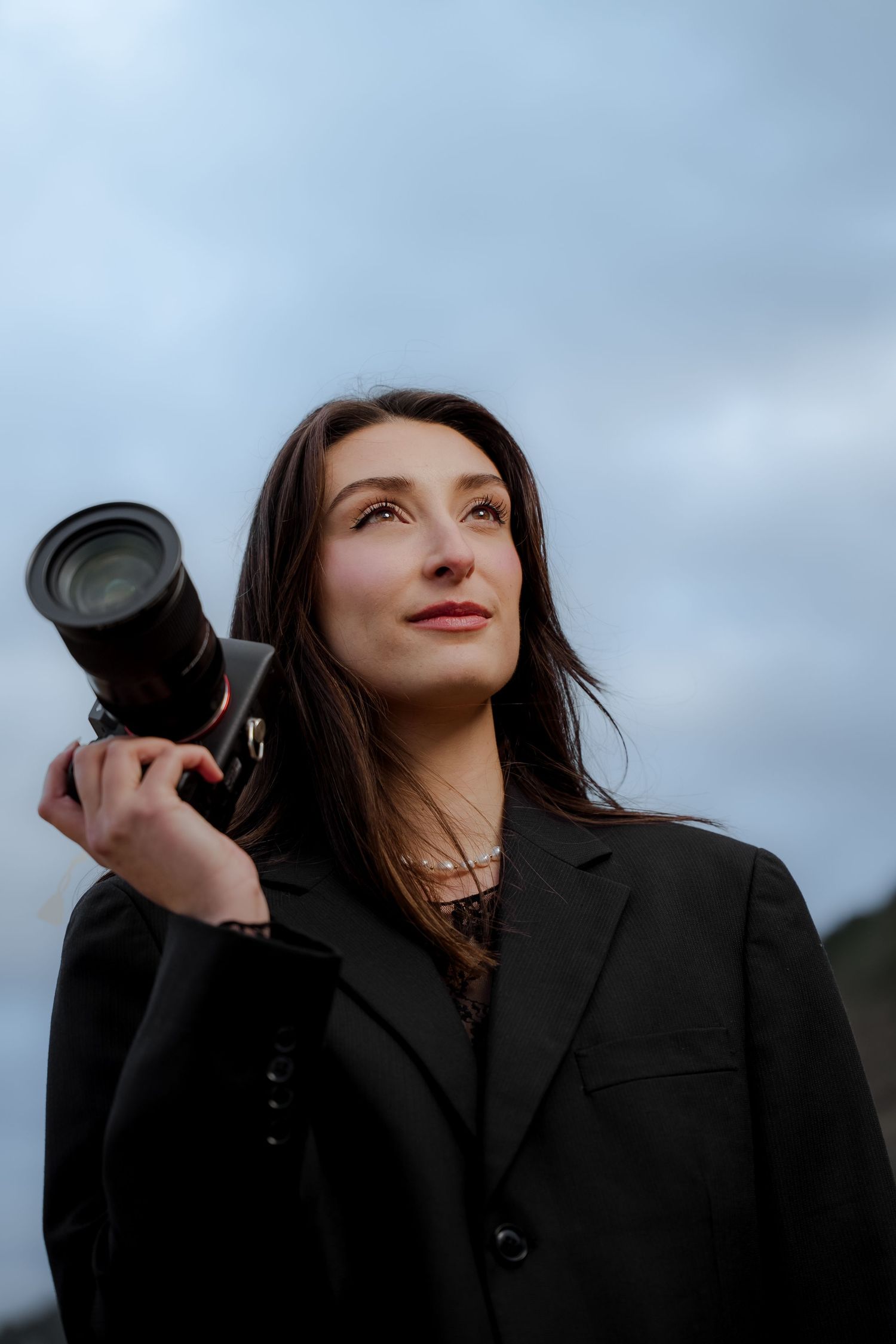 Woman in black blazer holding professional camera on shoulder, looking up at cloudy sky.