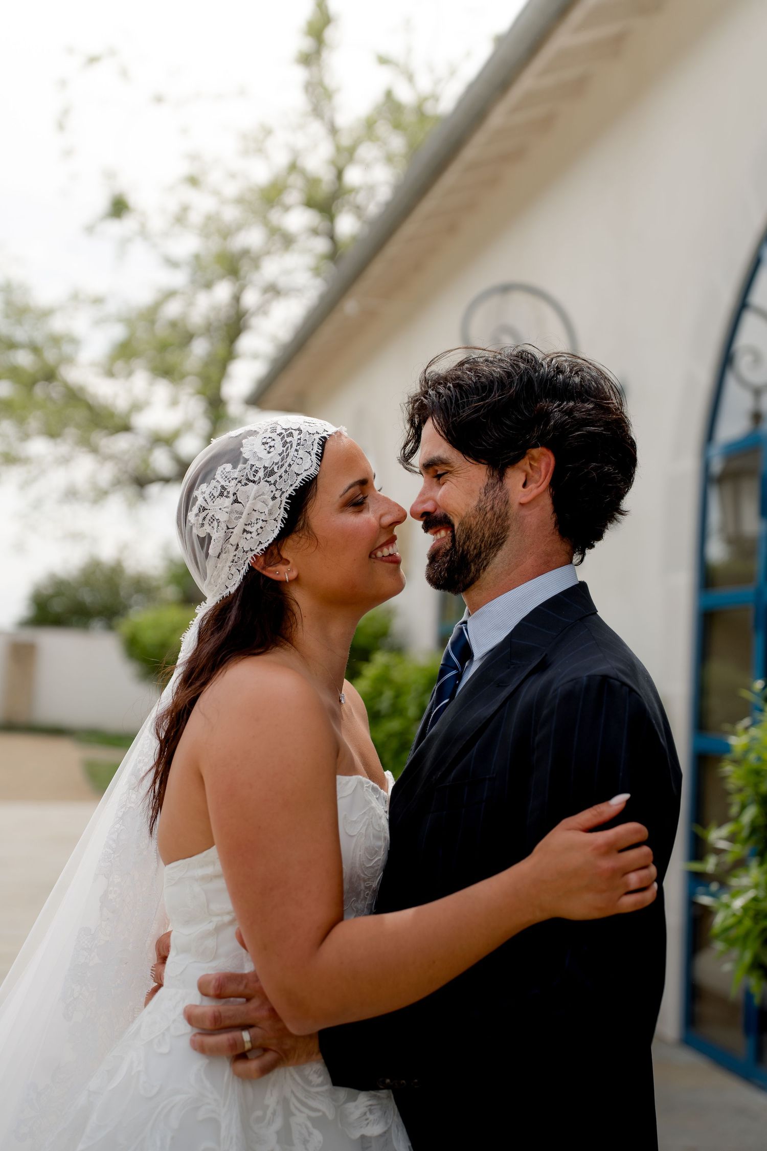 Smiling bride in lace veil and groom with beard embrace and gaze at each other outside white chapel.
