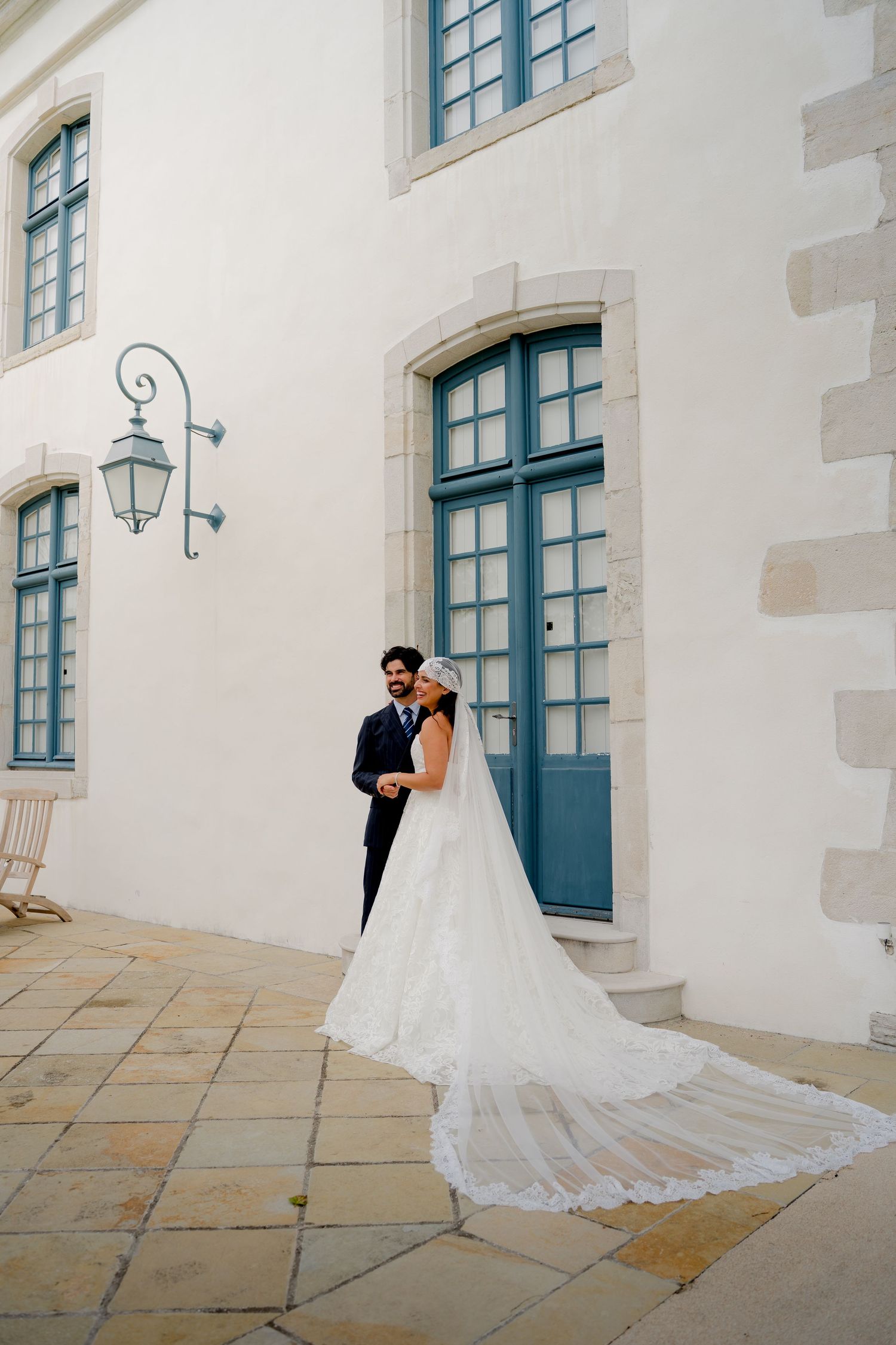 Bride in lace gown with cathedral veil stands with groom in navy suit outside elegant white French château.
