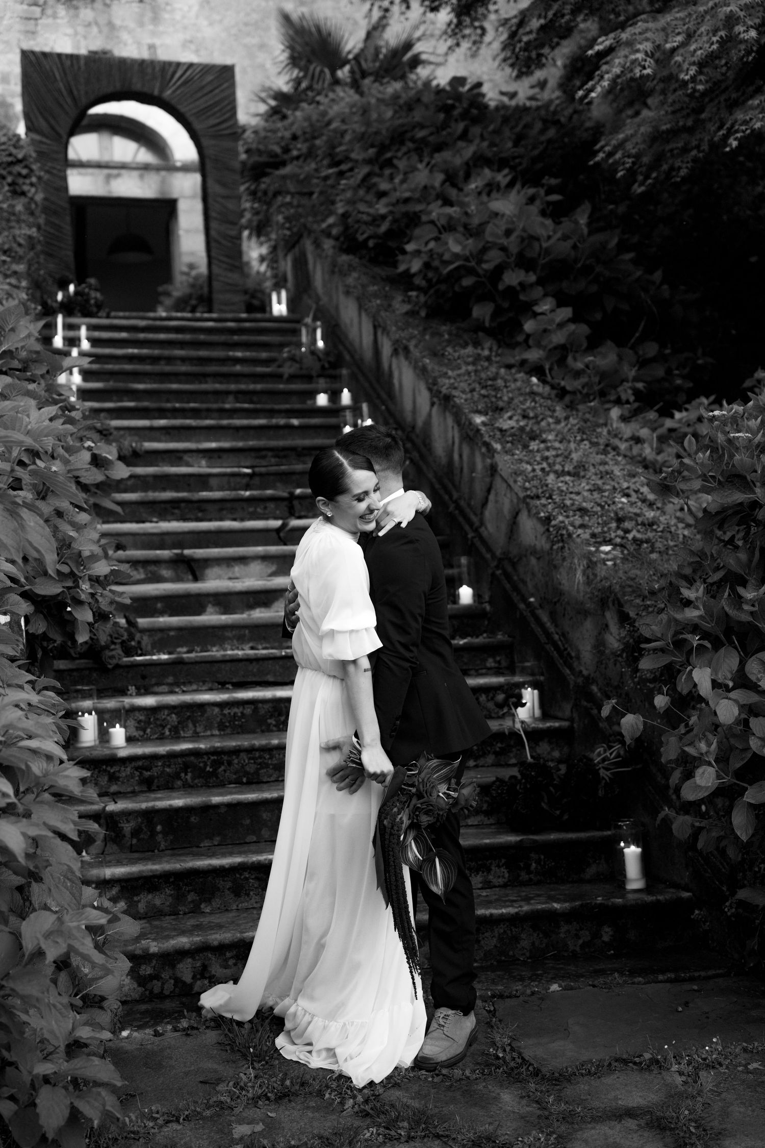 Black and white wedding portrait of a couple embracing at the foot of a grand stone staircase with candle decorations.