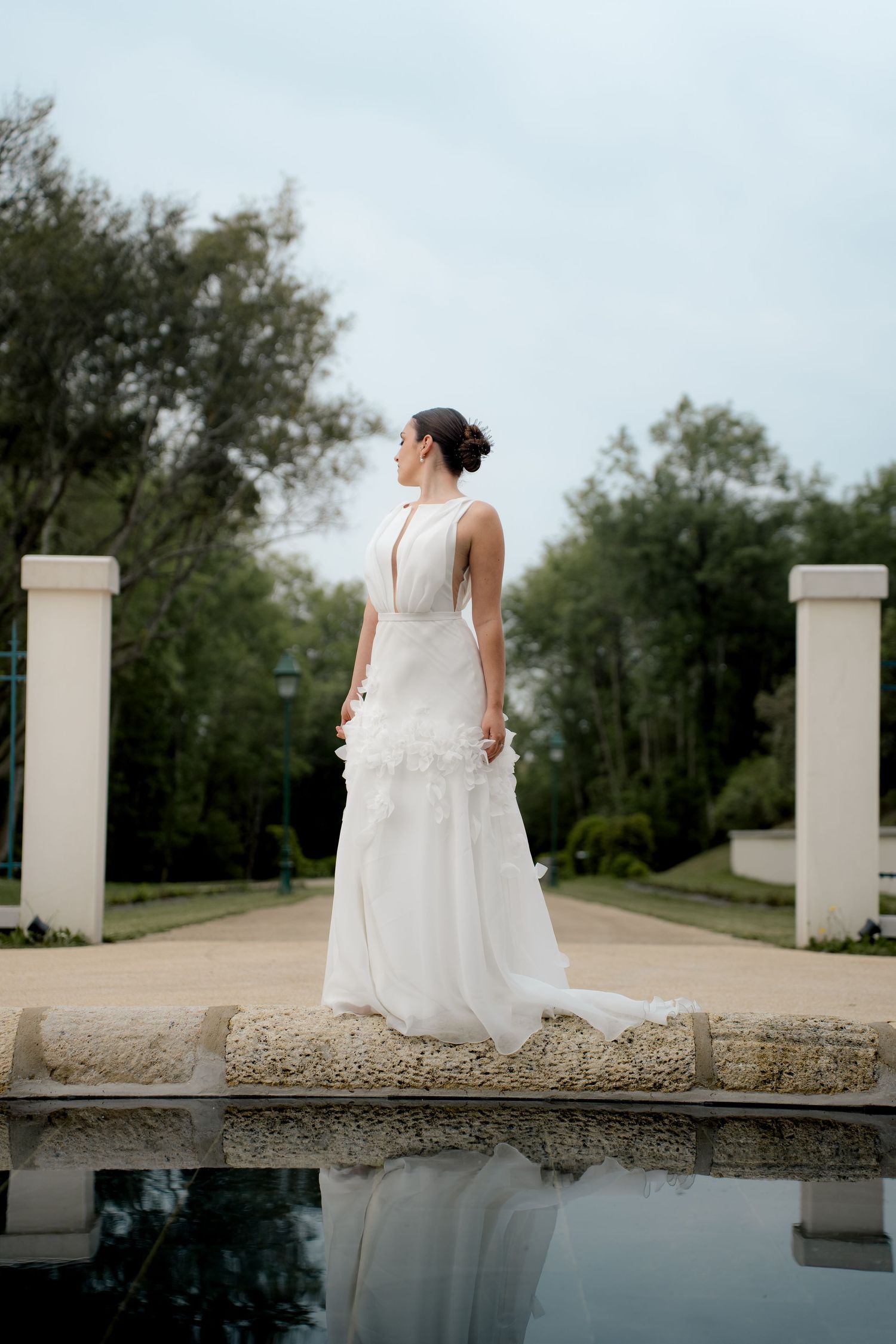 Elegant bride in white gown standing by reflective pool in lush garden.