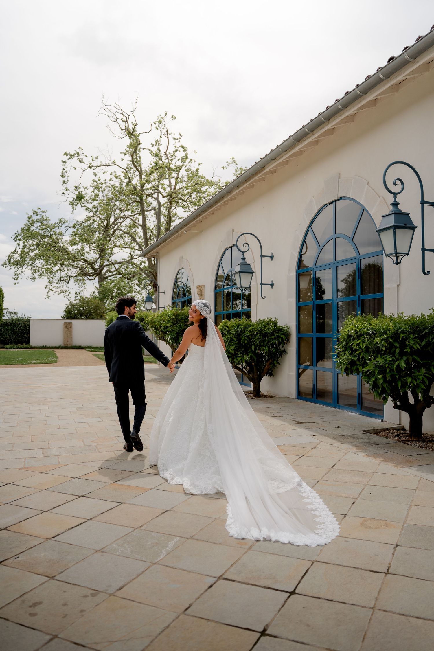 Bride in lace gown with long veil holds grooms hand outside elegant white venue with blue arched windows.