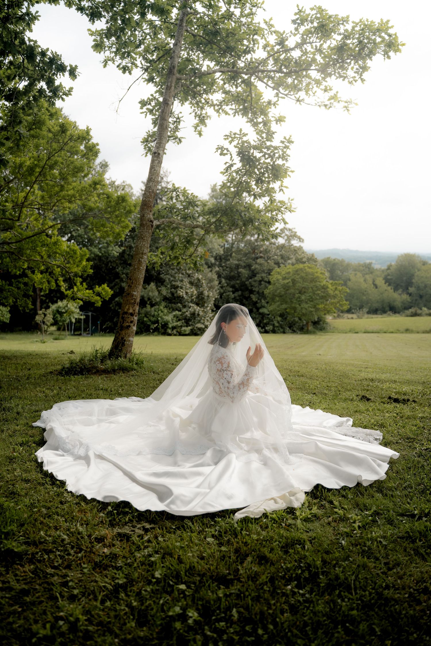 Bride in a flowing white lace wedding gown with a long veil sitting beneath a tree in a green field.