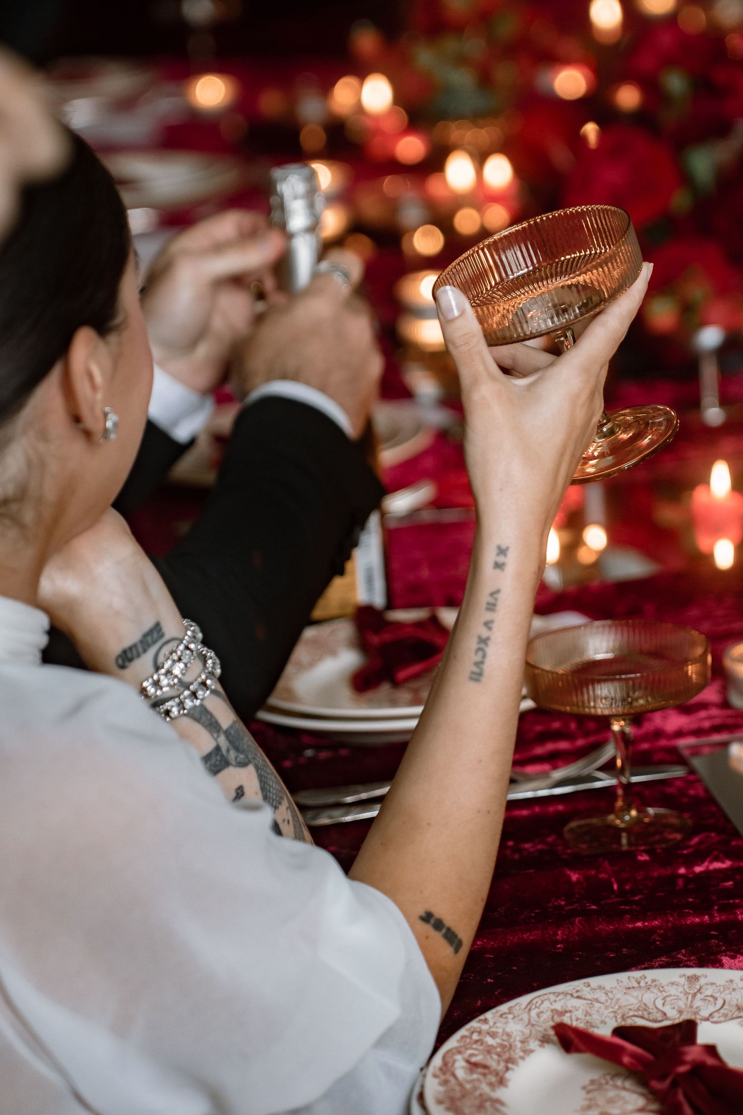 Tattooed bride raises amber cocktail glass at candlelit red velvet wedding reception table.