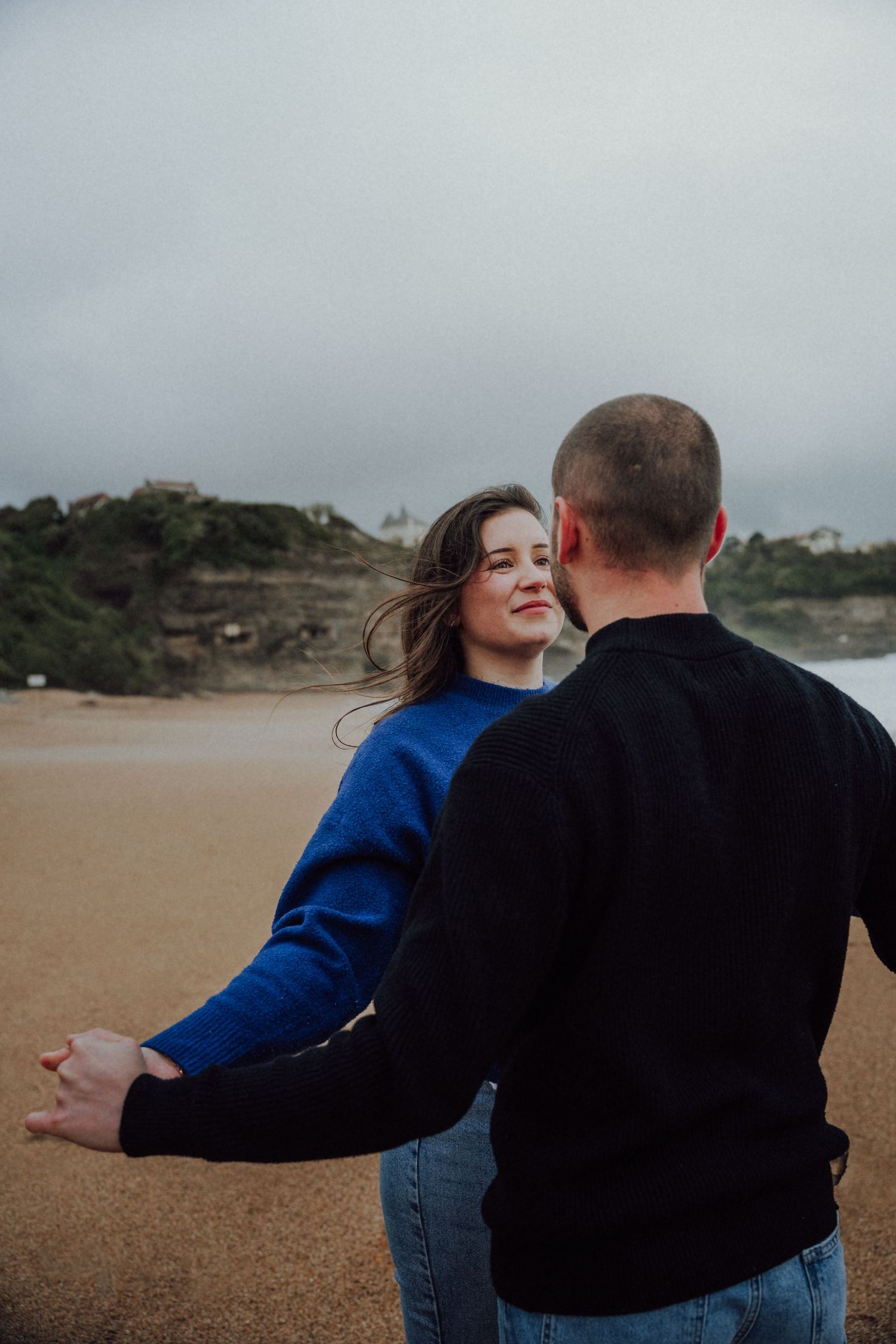 Couple dancing together on a windy beach, woman in blue sweater smiling while holding partners hands.