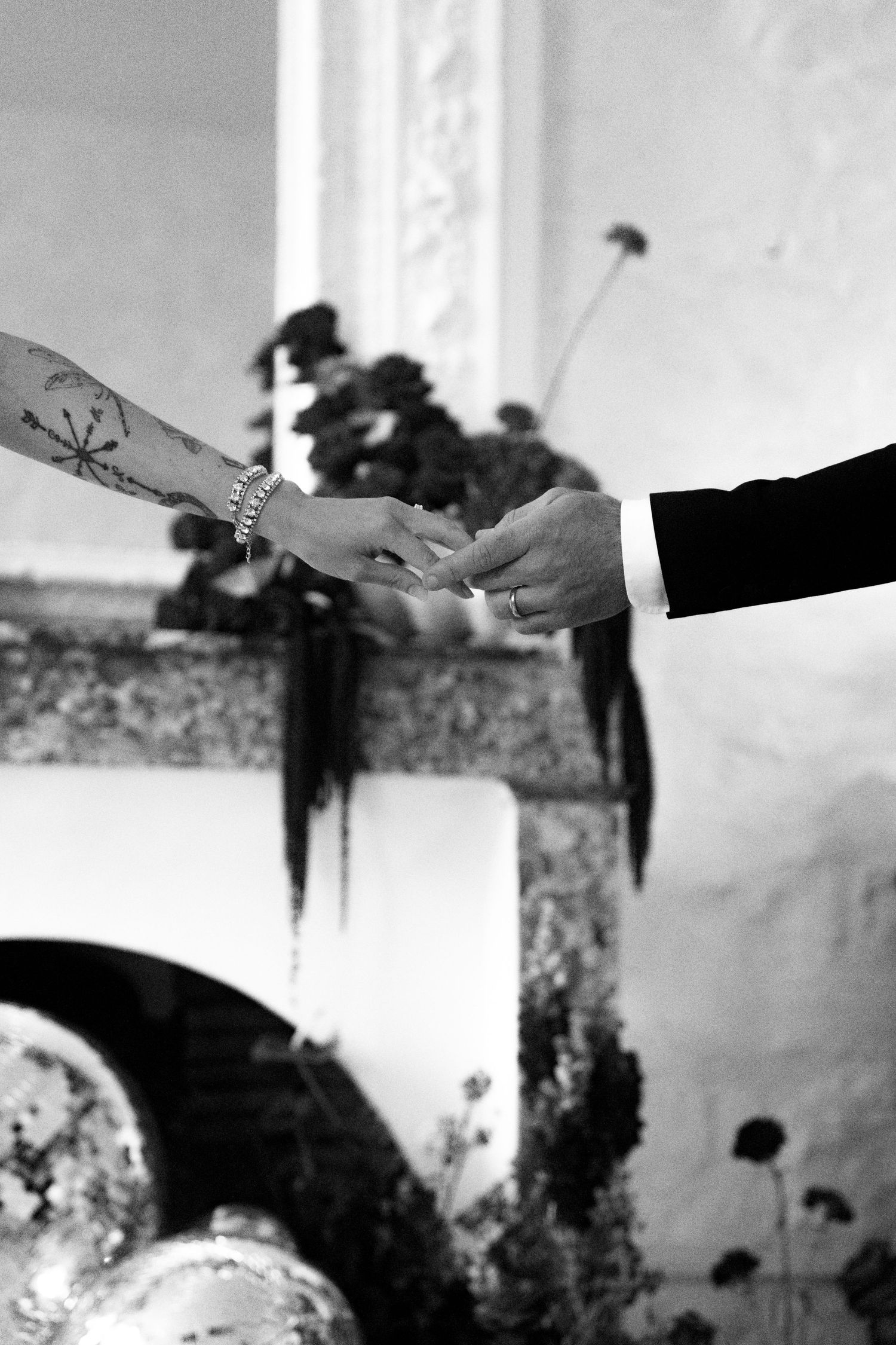 Groom placing wedding ring on tattooed bride's hand during intimate ceremony near floral fireplace.