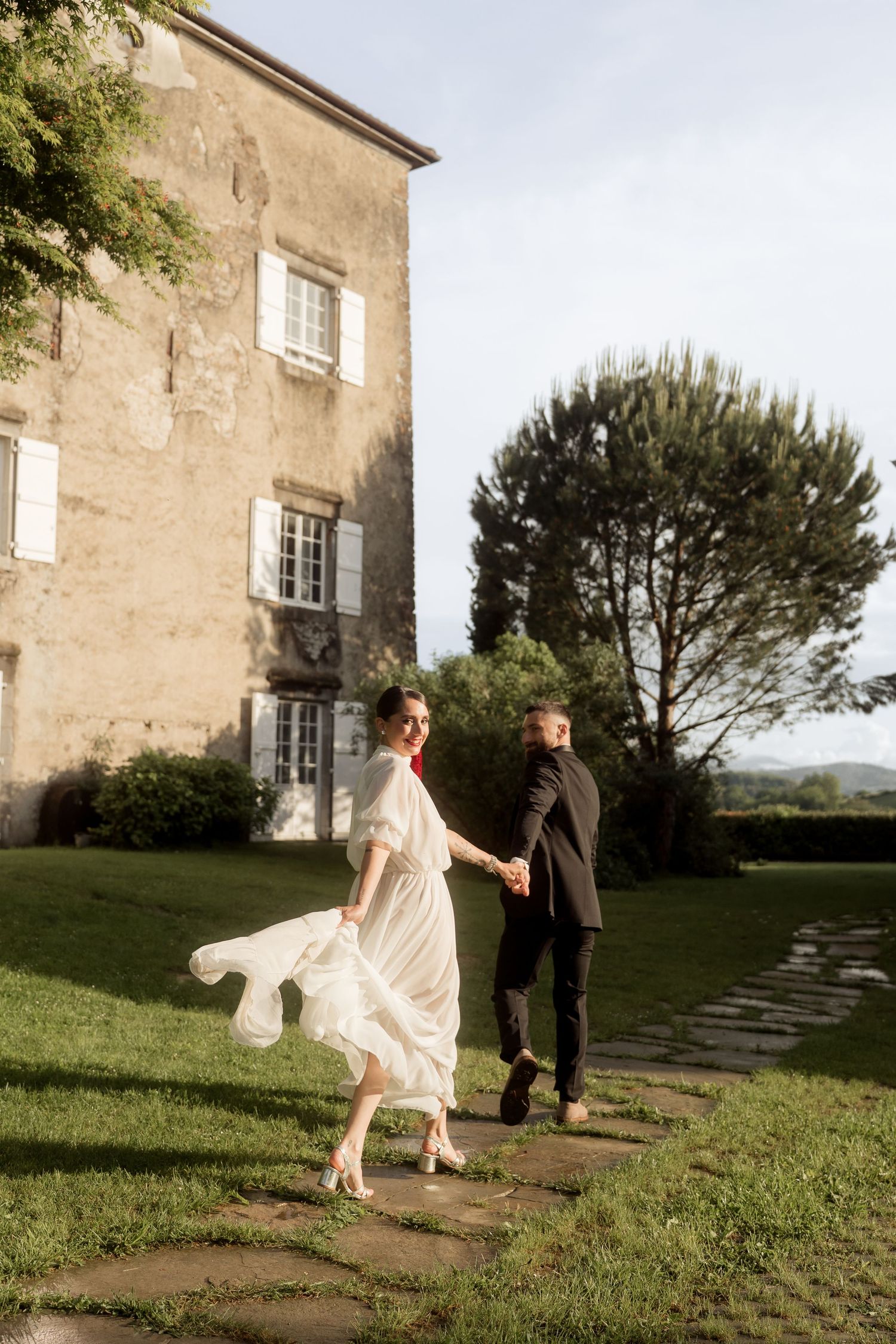 Bride in flowing white dress leads groom by hand along stone path beside rustic French chateau.