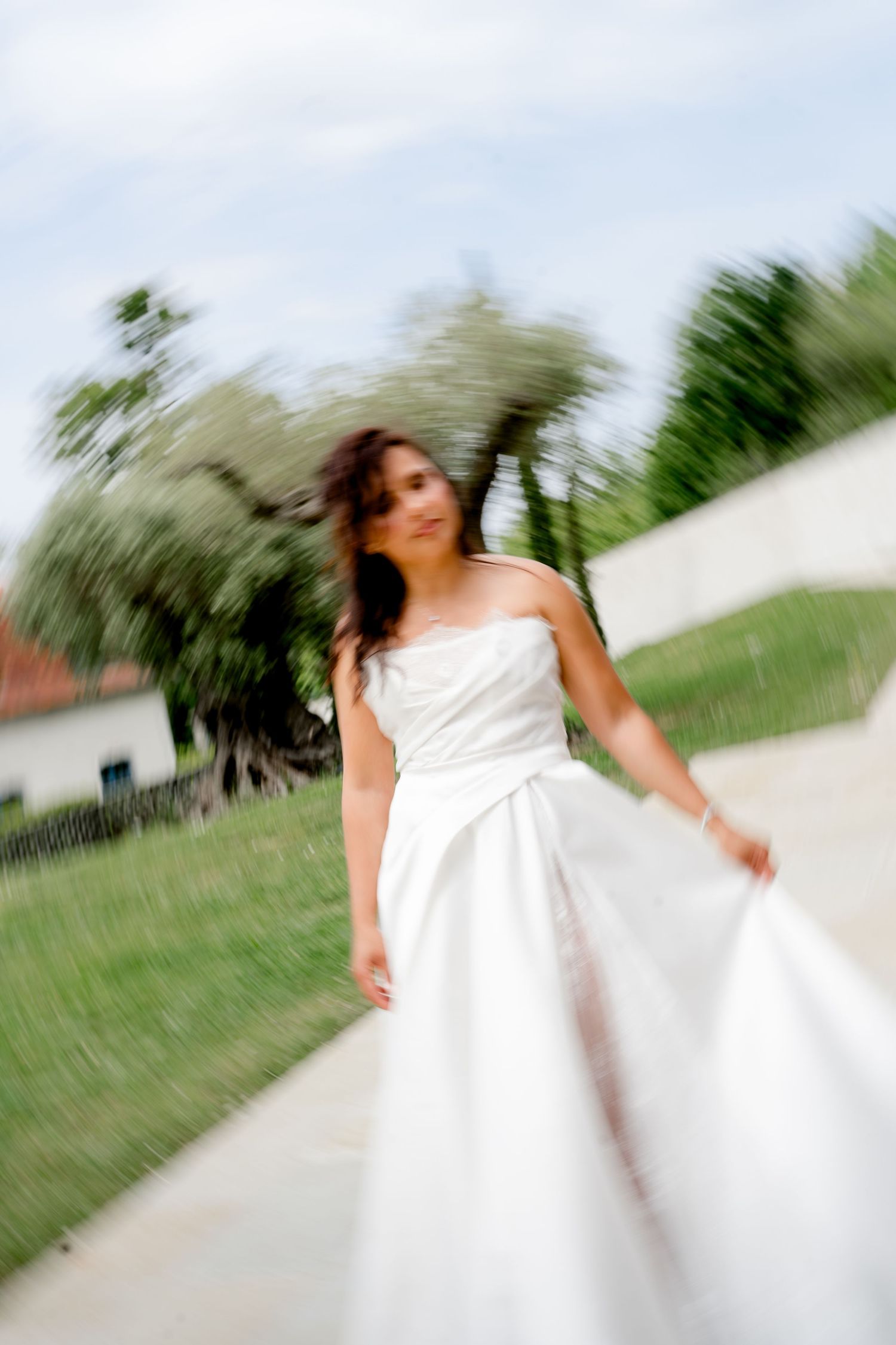 Motion-blurred artistic photo of a smiling bride in a white strapless gown twirling on a garden pathway.