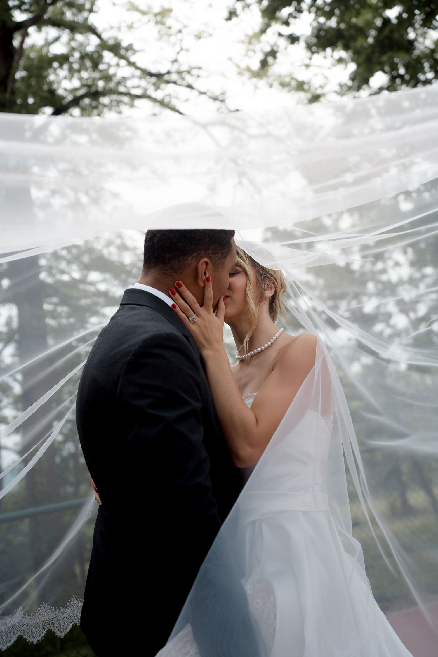 Bride and groom sharing a kiss under a flowing wedding veil outdoors beneath green trees.
