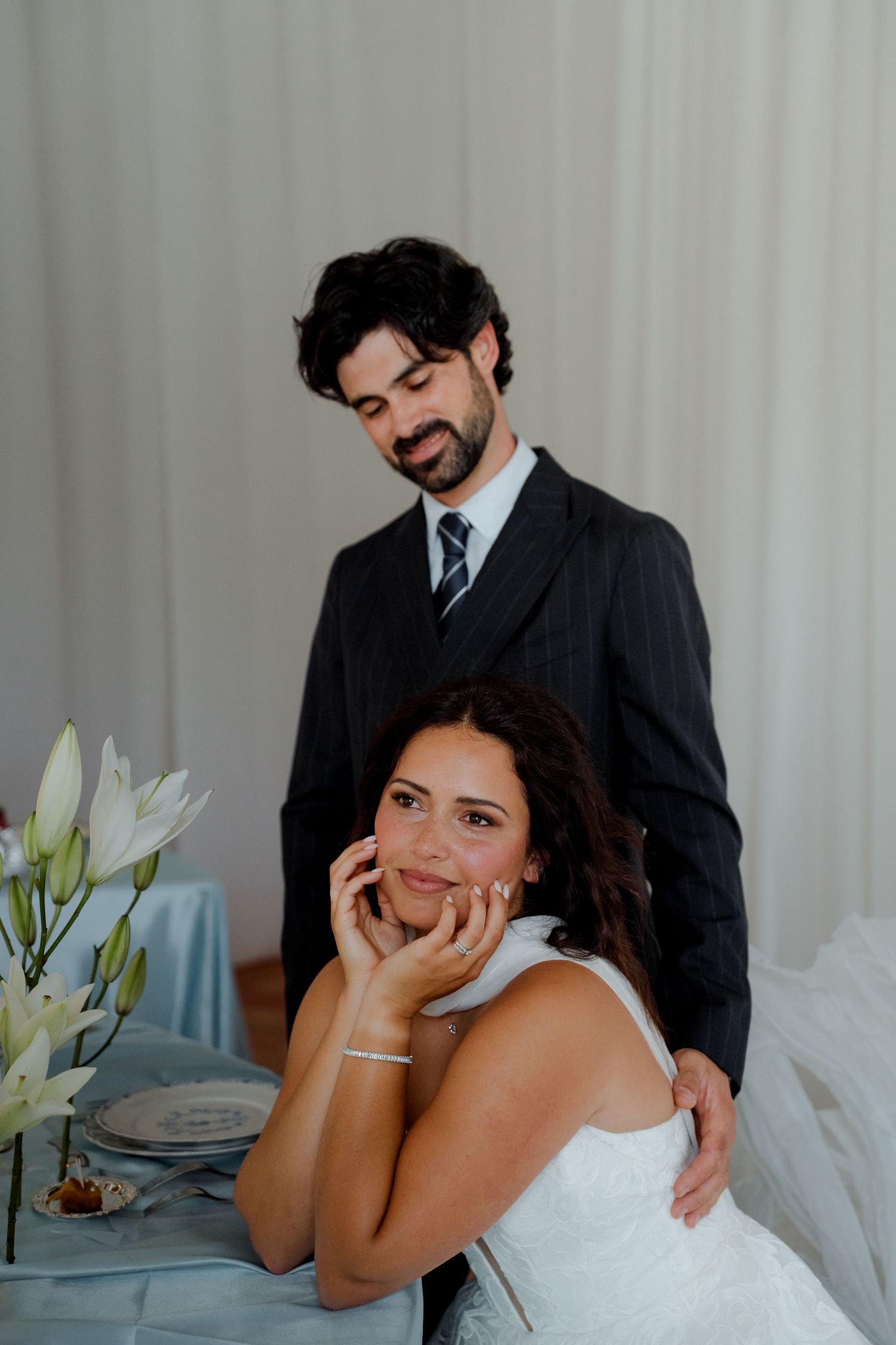 Bride resting her face in her hands while groom stands behind her at reception table.