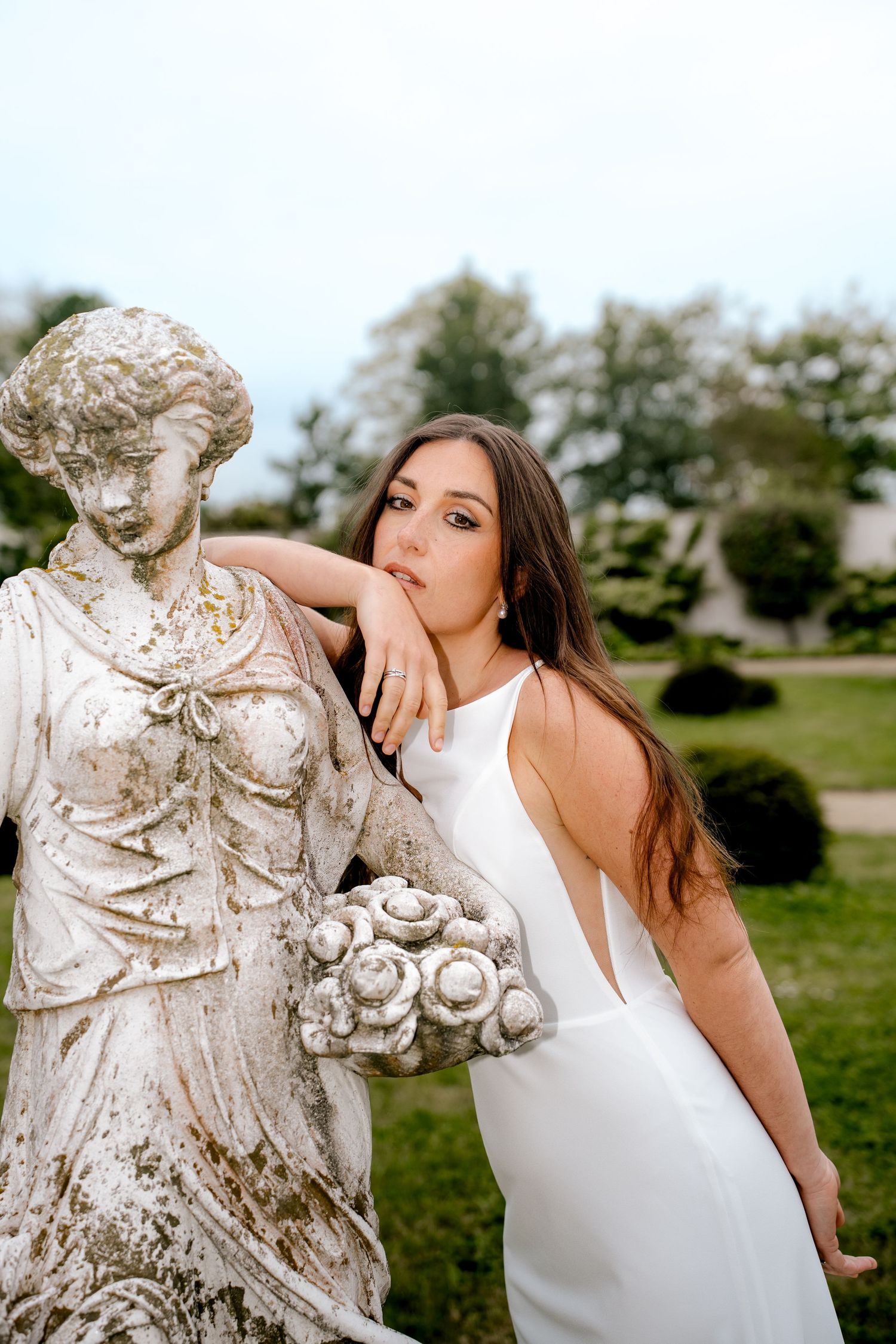 Brunette bride in sleek white gown posing beside weathered classical stone statue in garden.