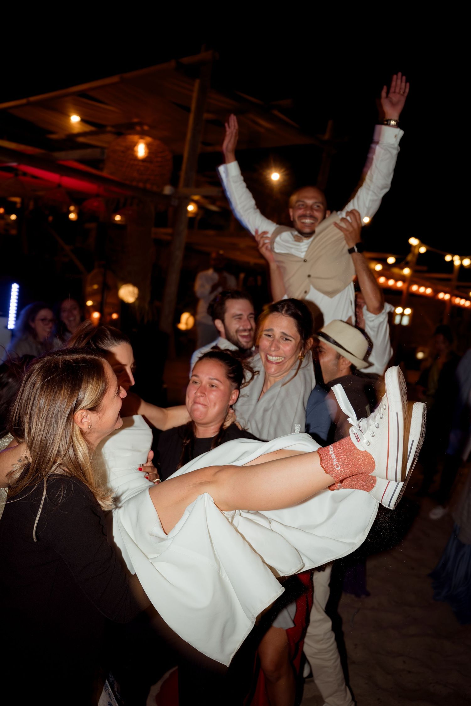 Bride being lifted by cheering wedding guests during lively beach reception.