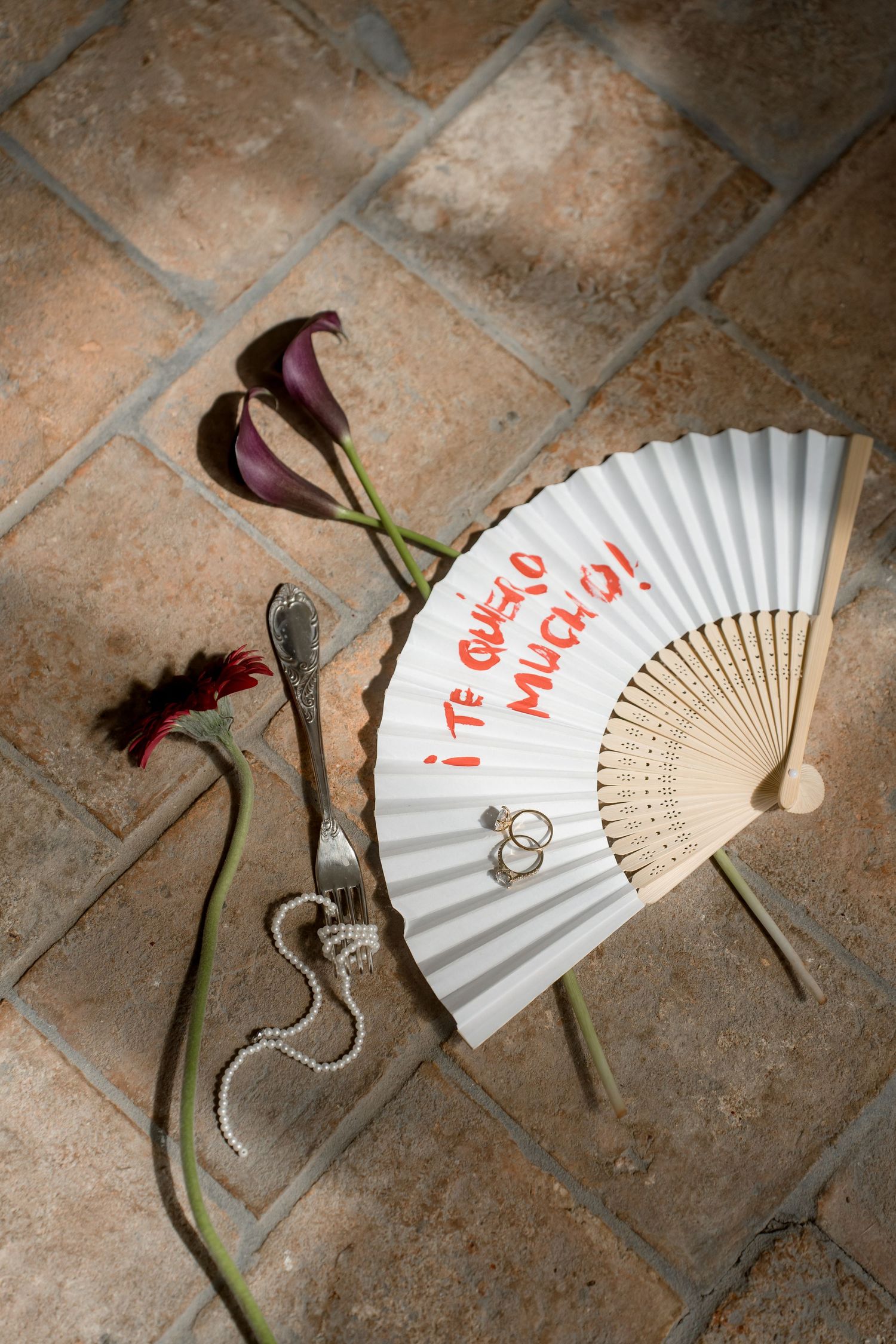White hand fan with 'Te Quiero Mucho' message, wedding rings, pearl necklace, and dark calla lilies on terracotta tiles.