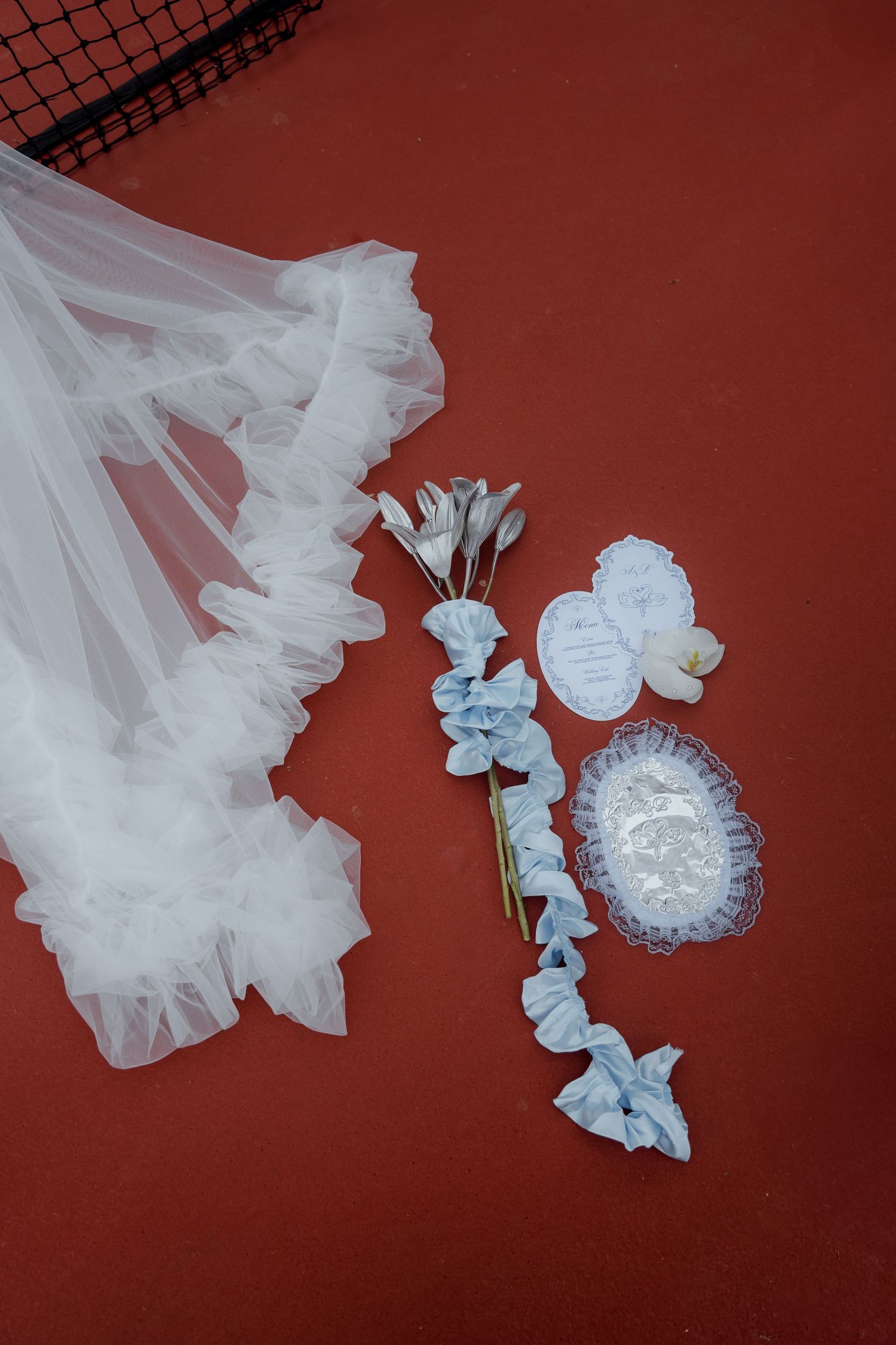 White ruffled wedding veil beside blue fabric flower bouquet and lace accessories on red court surface.