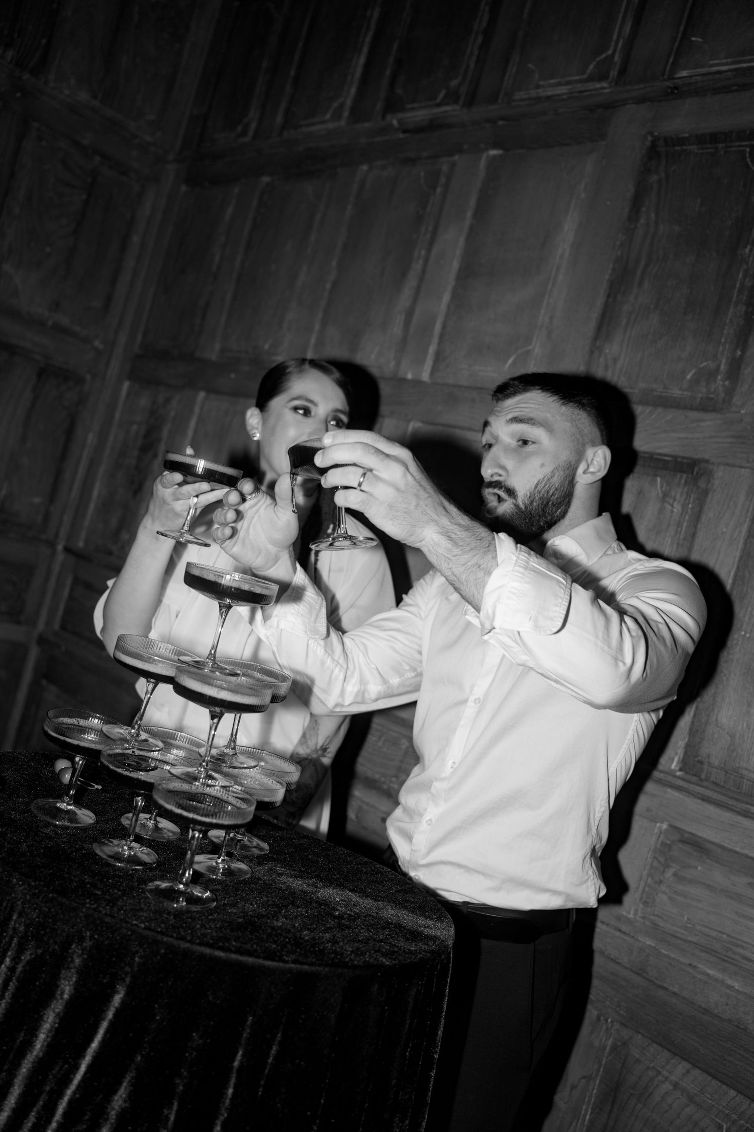 Black and white photo of couple playfully pouring champagne tower at wedding reception.