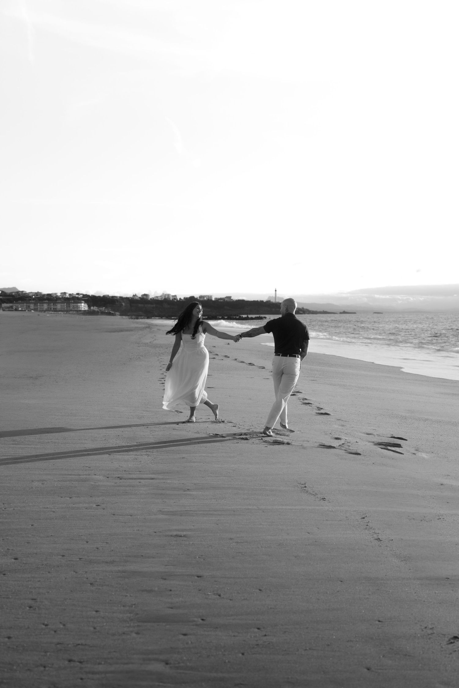 Black and white photo of a couple holding hands and walking along a sandy beach at sunset.