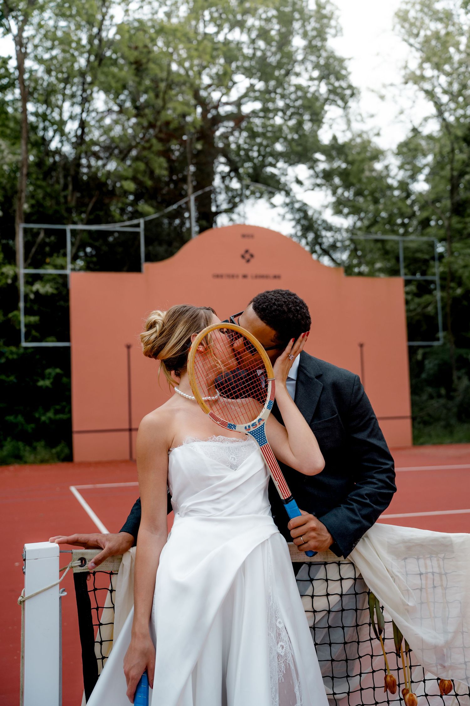 Bride and groom kissing behind a tennis racket at the net on a red clay court.