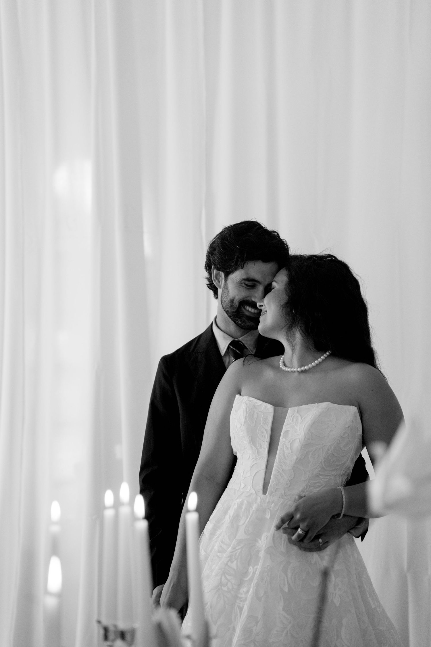 Black and white portrait of smiling bride in strapless gown and groom embracing near candlelit drapes.