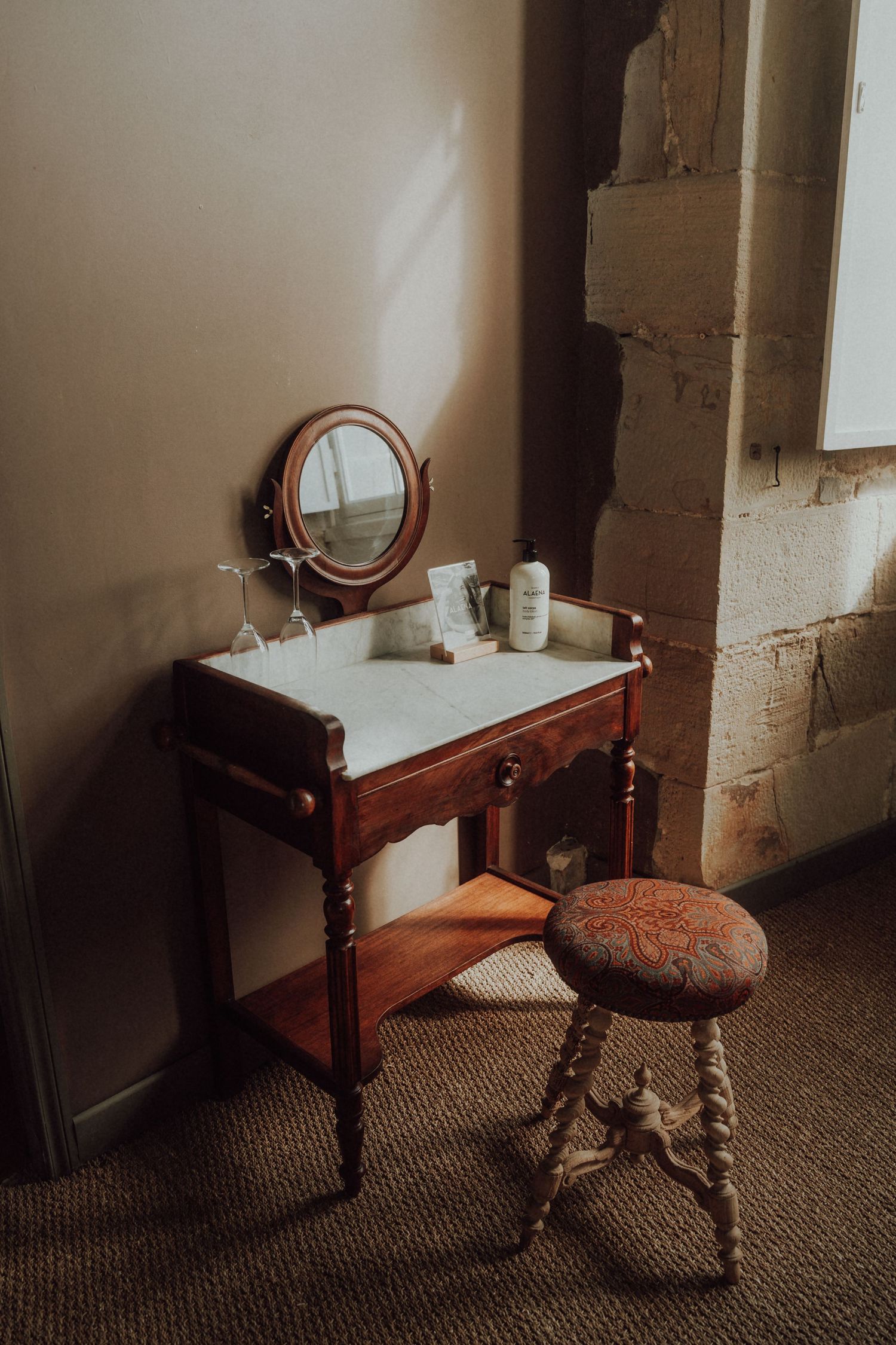 Antique wooden vanity table with marble top, round mirror, and ornate stool in sunlit rustic room.