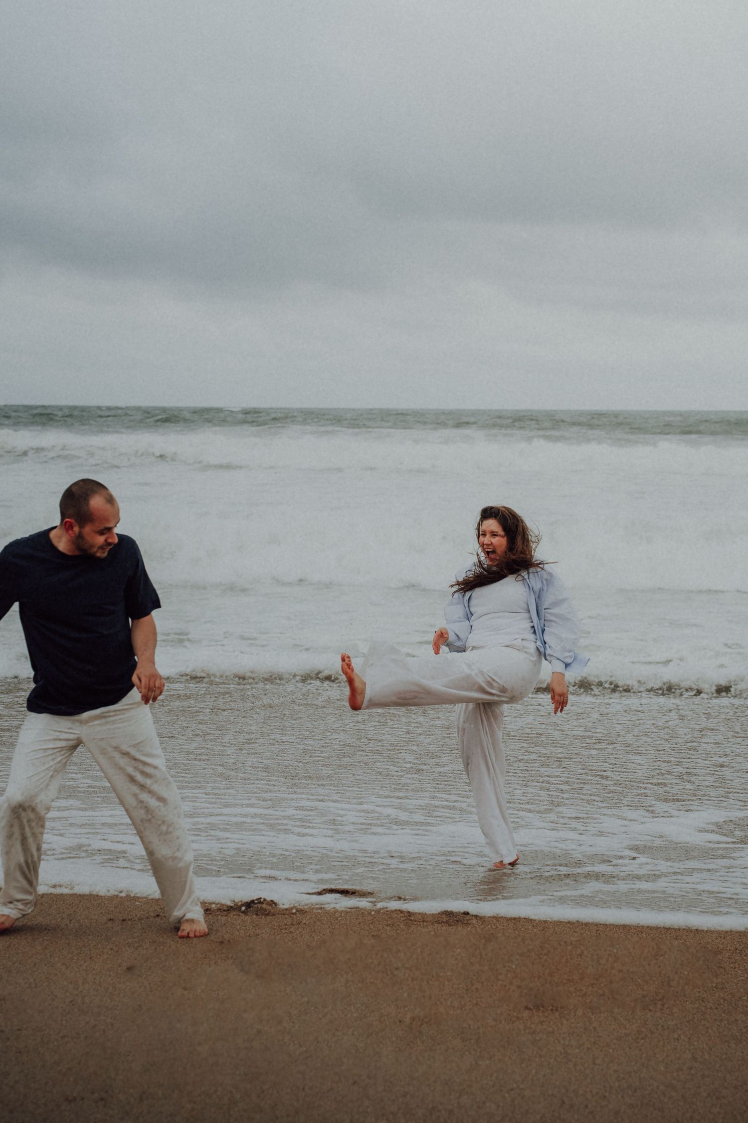 Playful couple on a cloudy beach, woman kicking leg up while man watches and laughs near ocean waves.