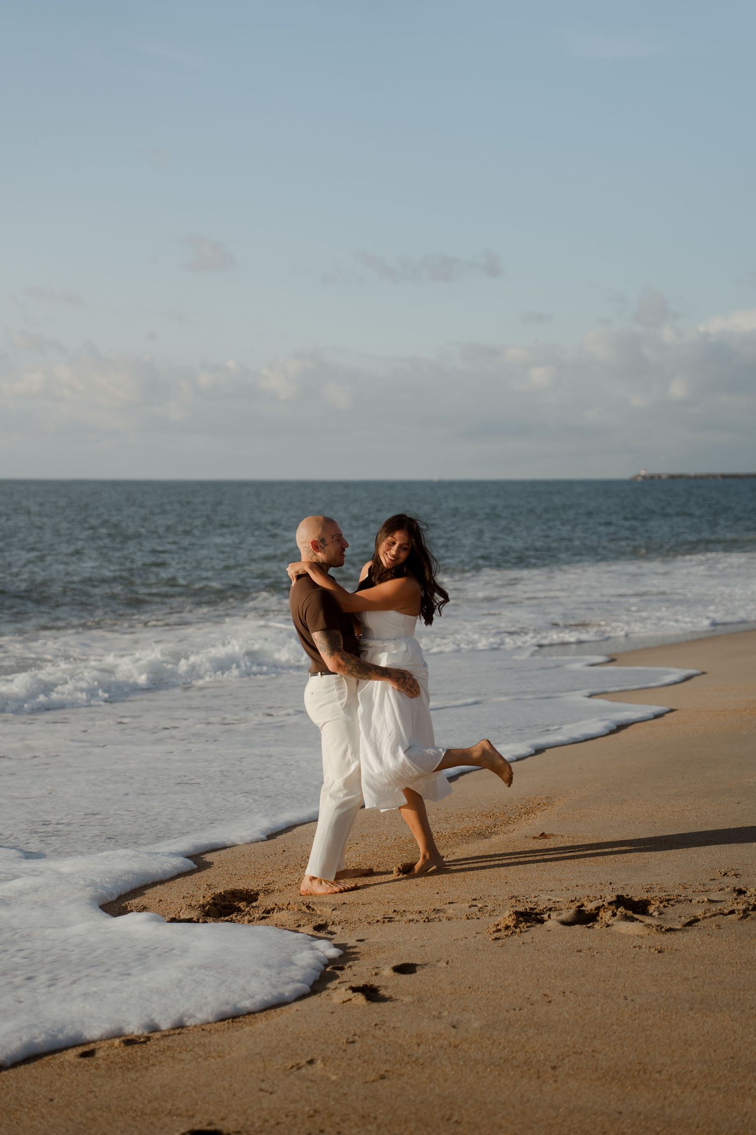 A bald tattooed man lifts a woman in a white dress as waves wash ashore during a beach photo session.