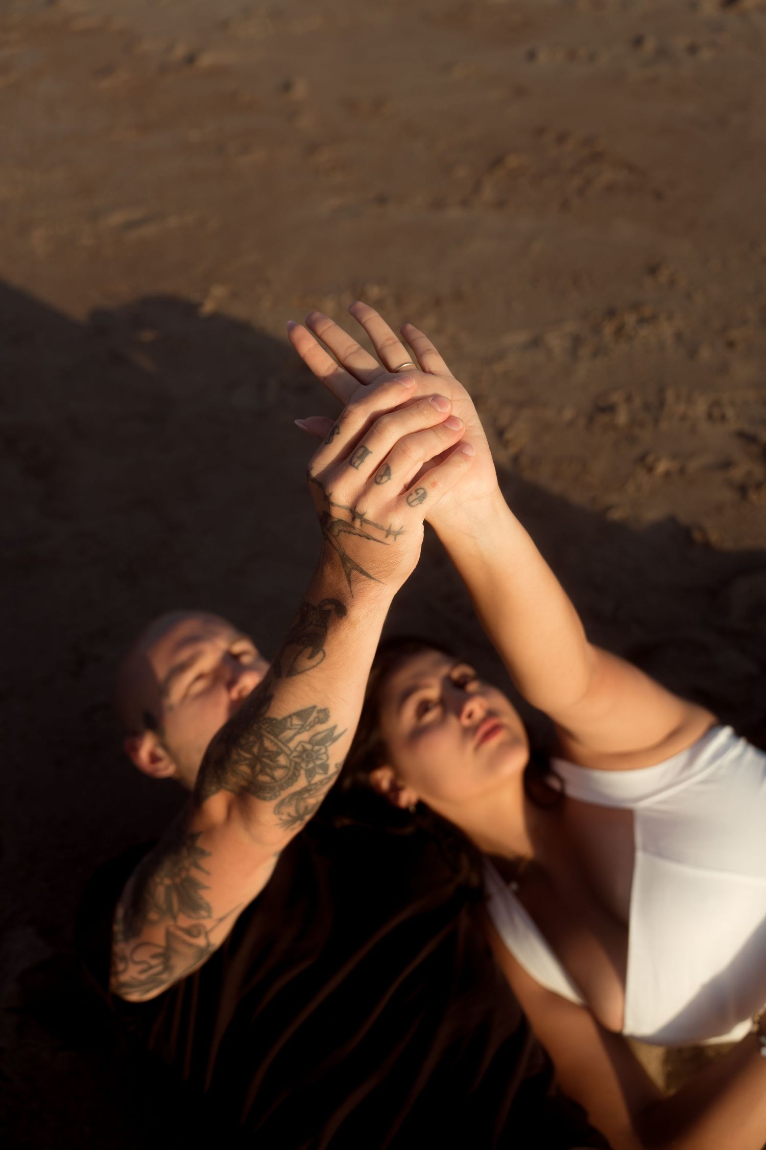 Couple lying on beach sand holding hands up toward sky, man with tattooed arm, golden light.