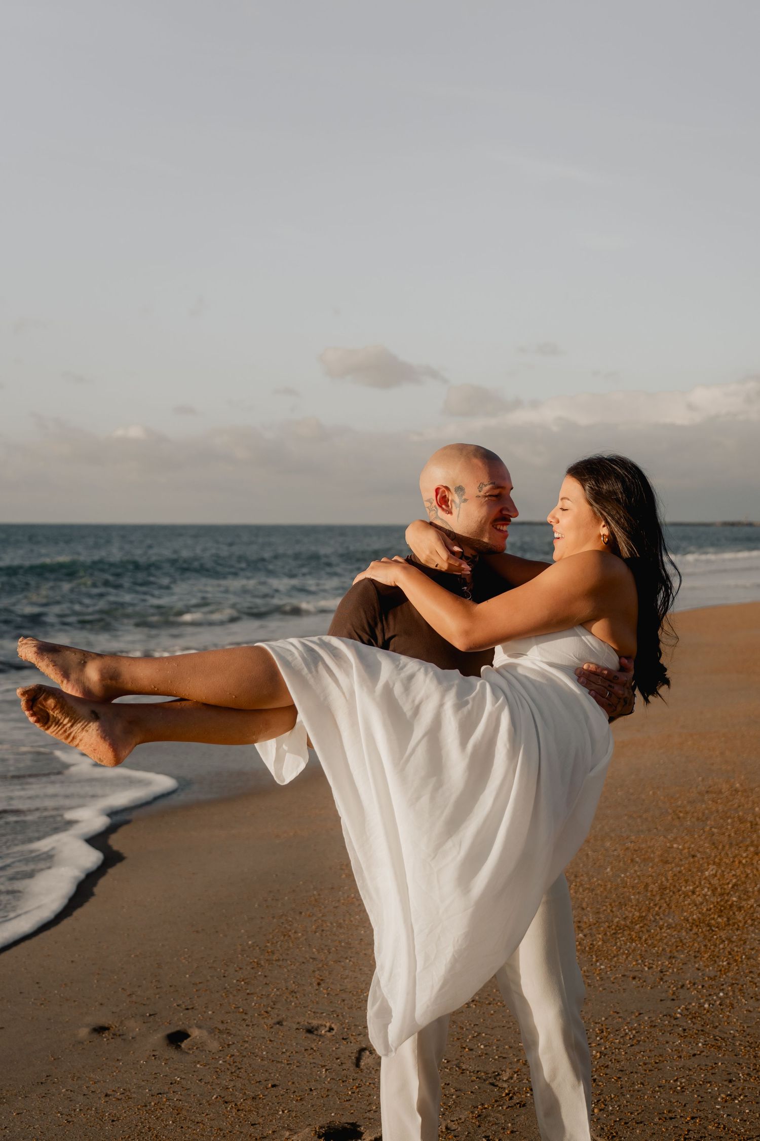 A man carries a woman in a white dress in his arms on a sunny beach with ocean waves behind them.