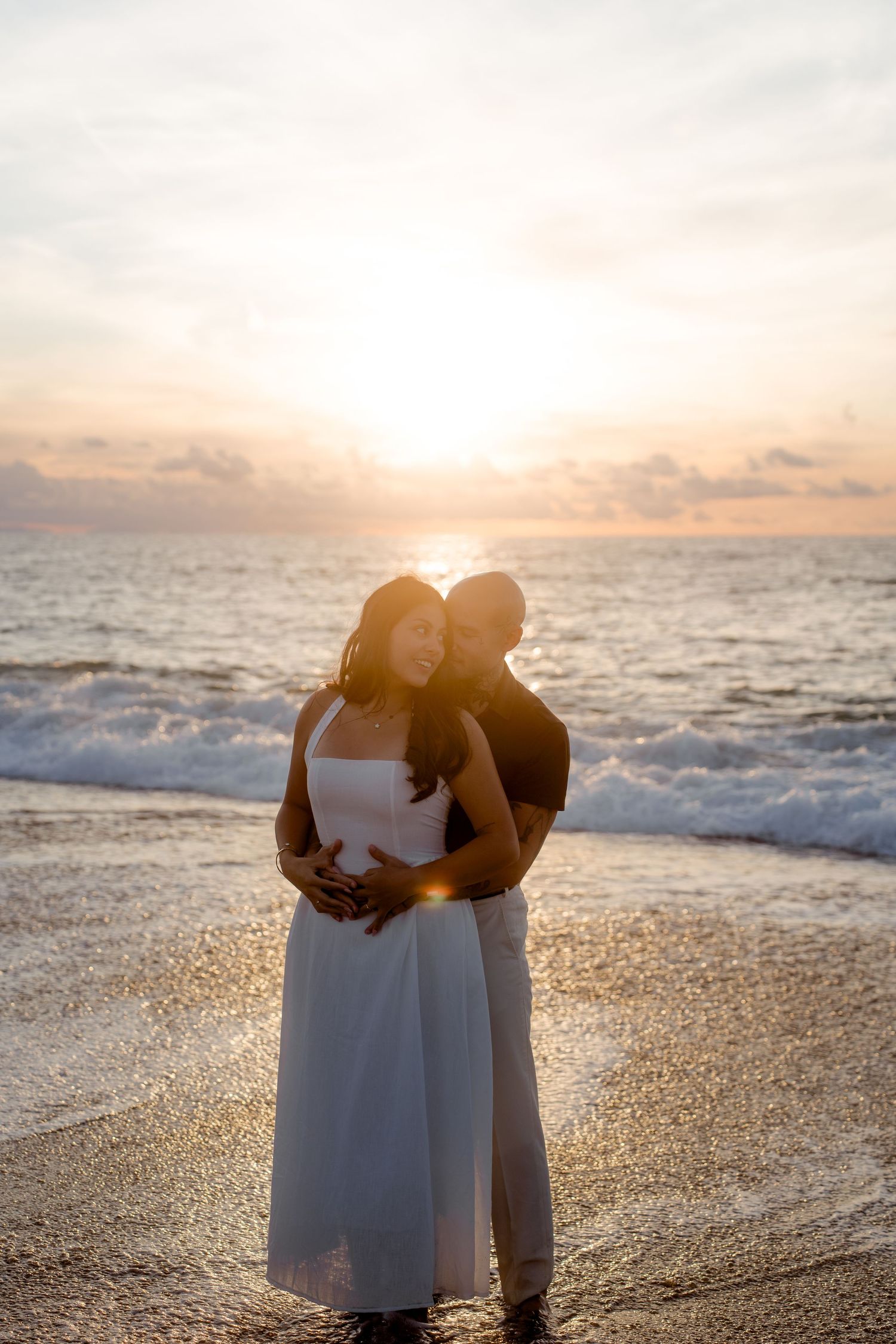 Couple embracing on beach at golden sunset, woman in white dress, waves crashing behind them.
