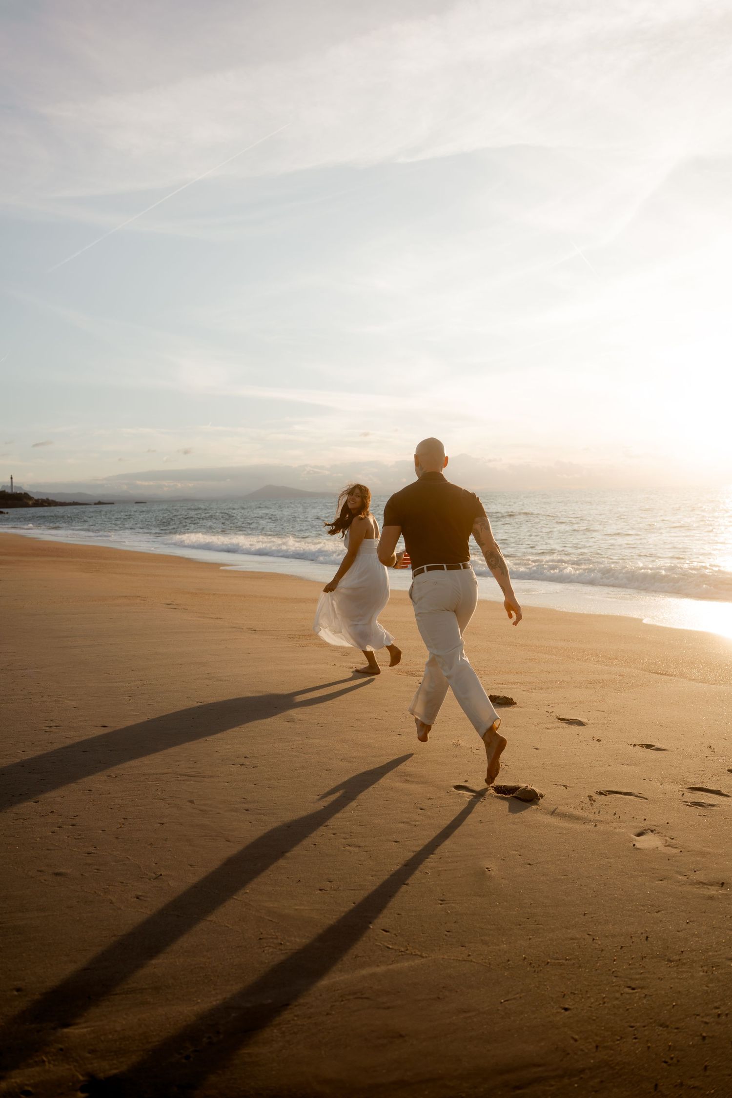 Couple running hand-in-hand barefoot on a sandy beach during golden hour sunset, casting long shadows.