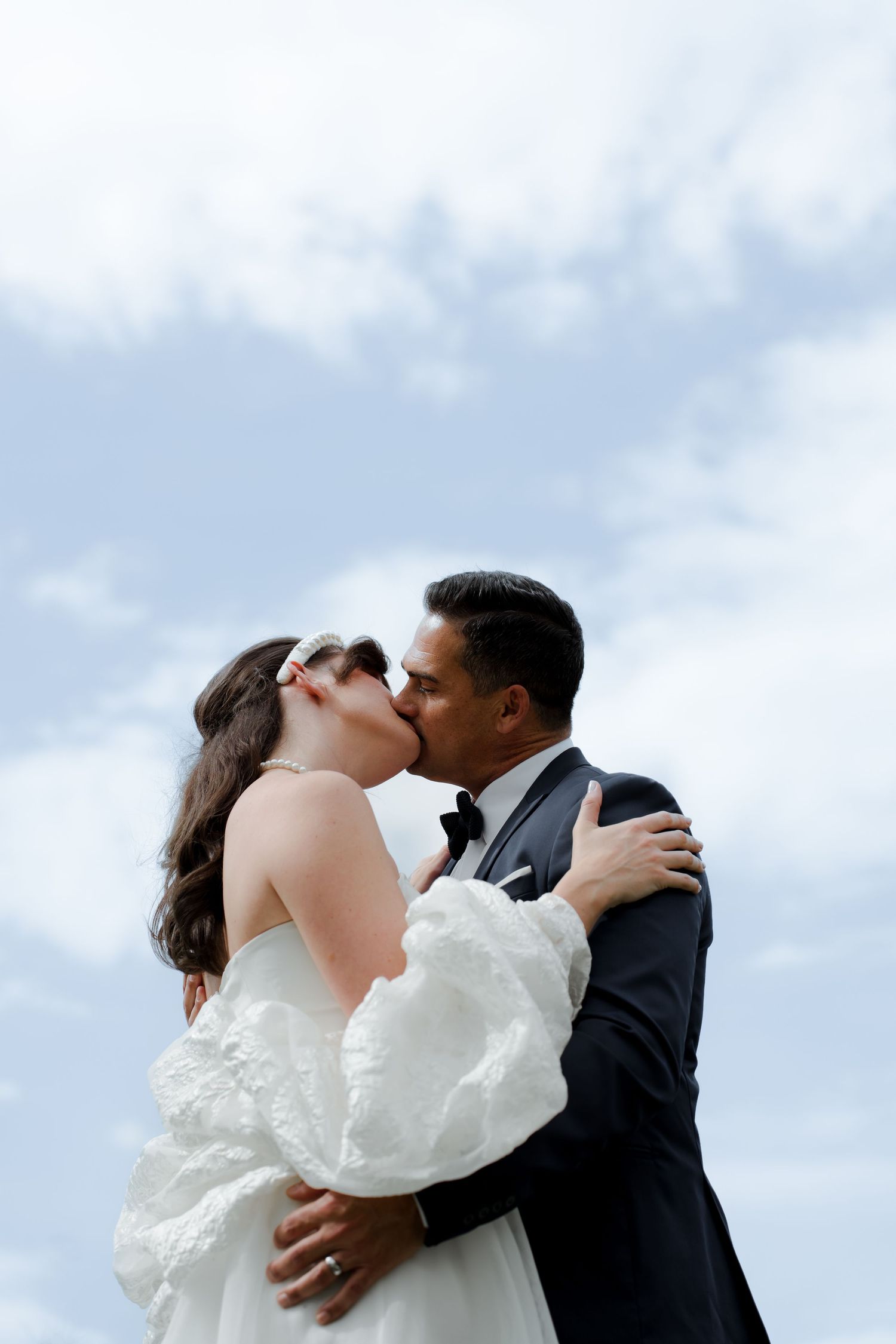 Bride and groom share a passionate kiss against a cloudy blue sky, bride wearing white ruffled sleeves.