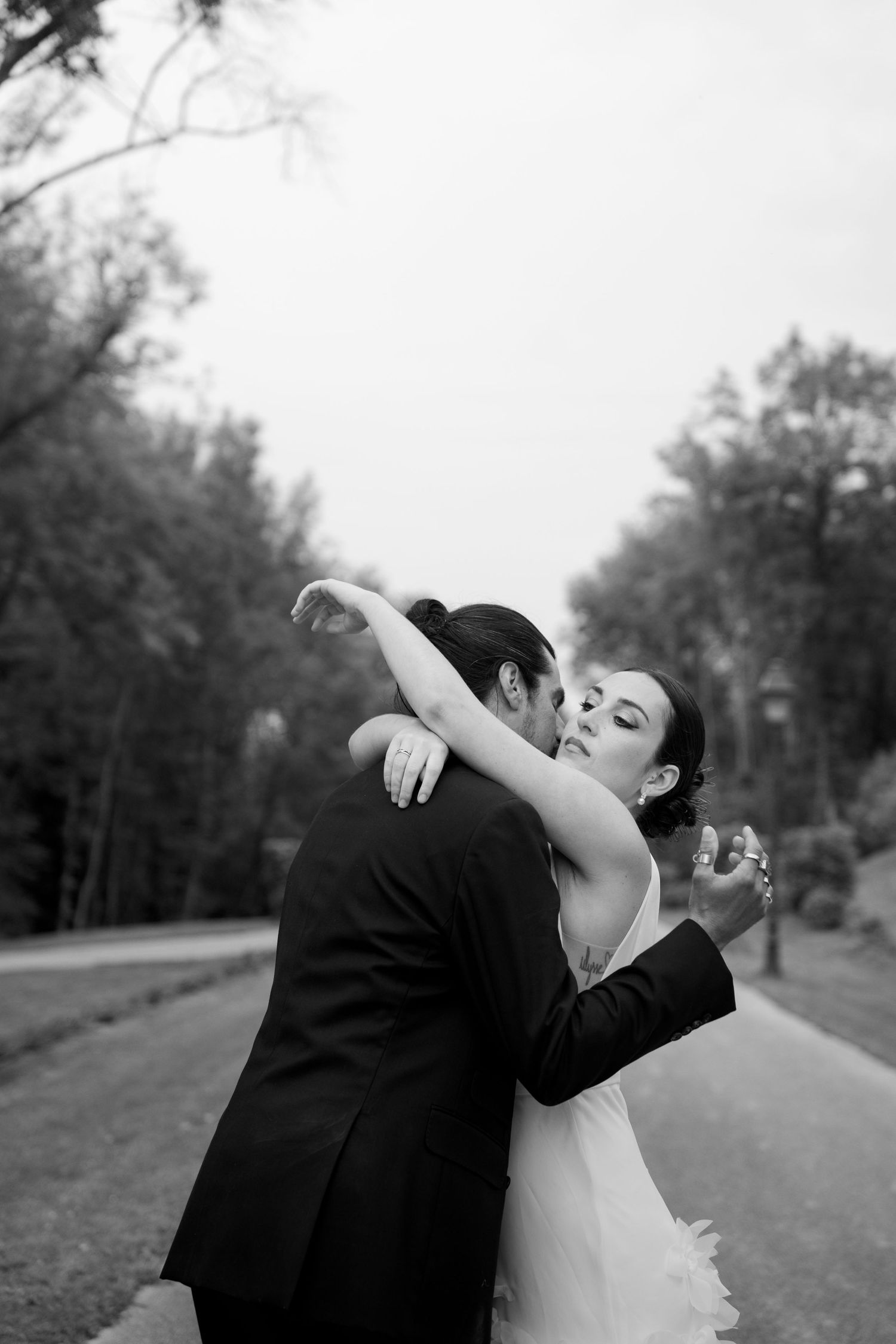 Black and white photo of bride and groom sharing romantic kiss on tree-lined path outdoors.