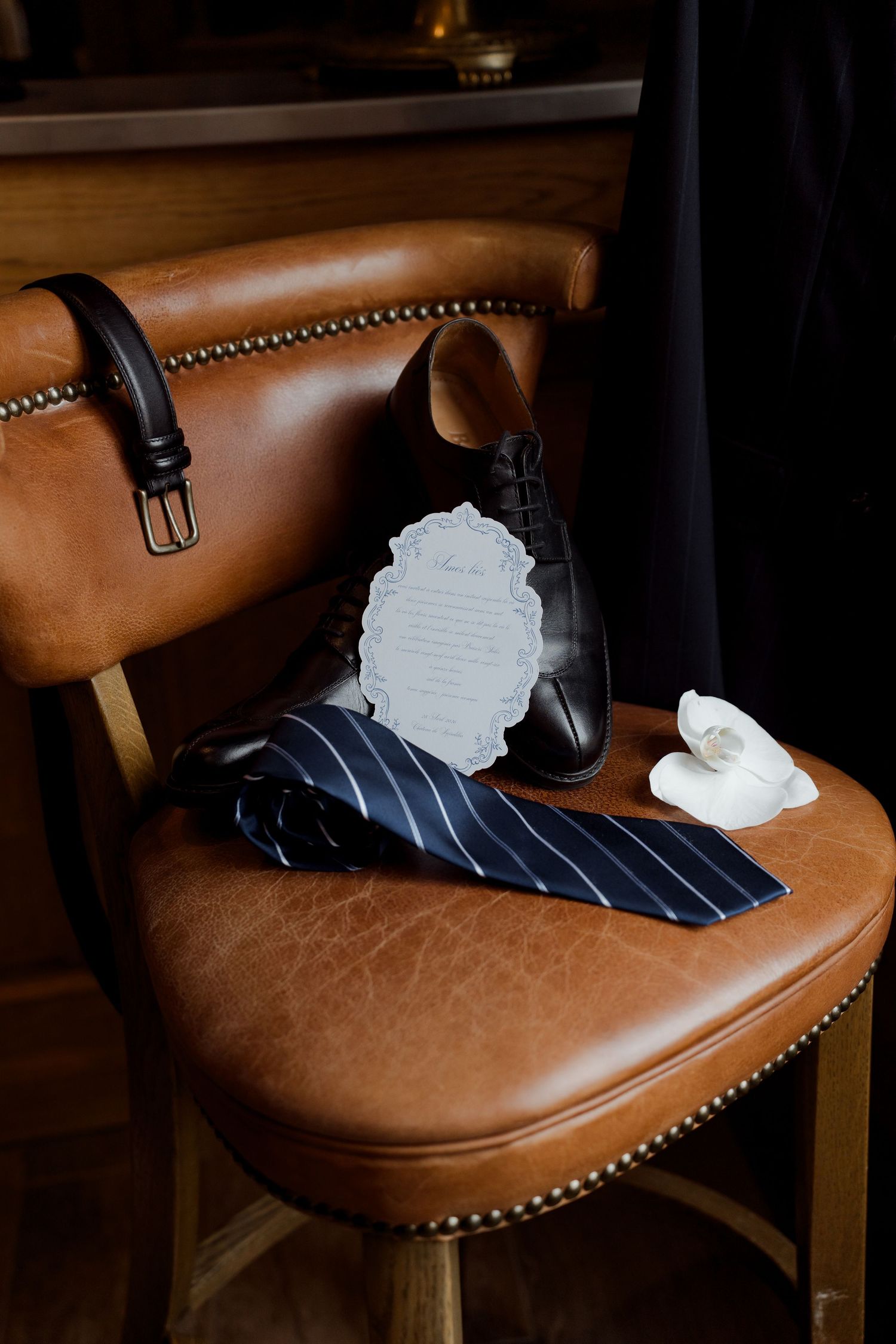 Groom wedding details with black dress shoes, striped navy tie, leather belt, boutonniere, and wedding vow card on chair.