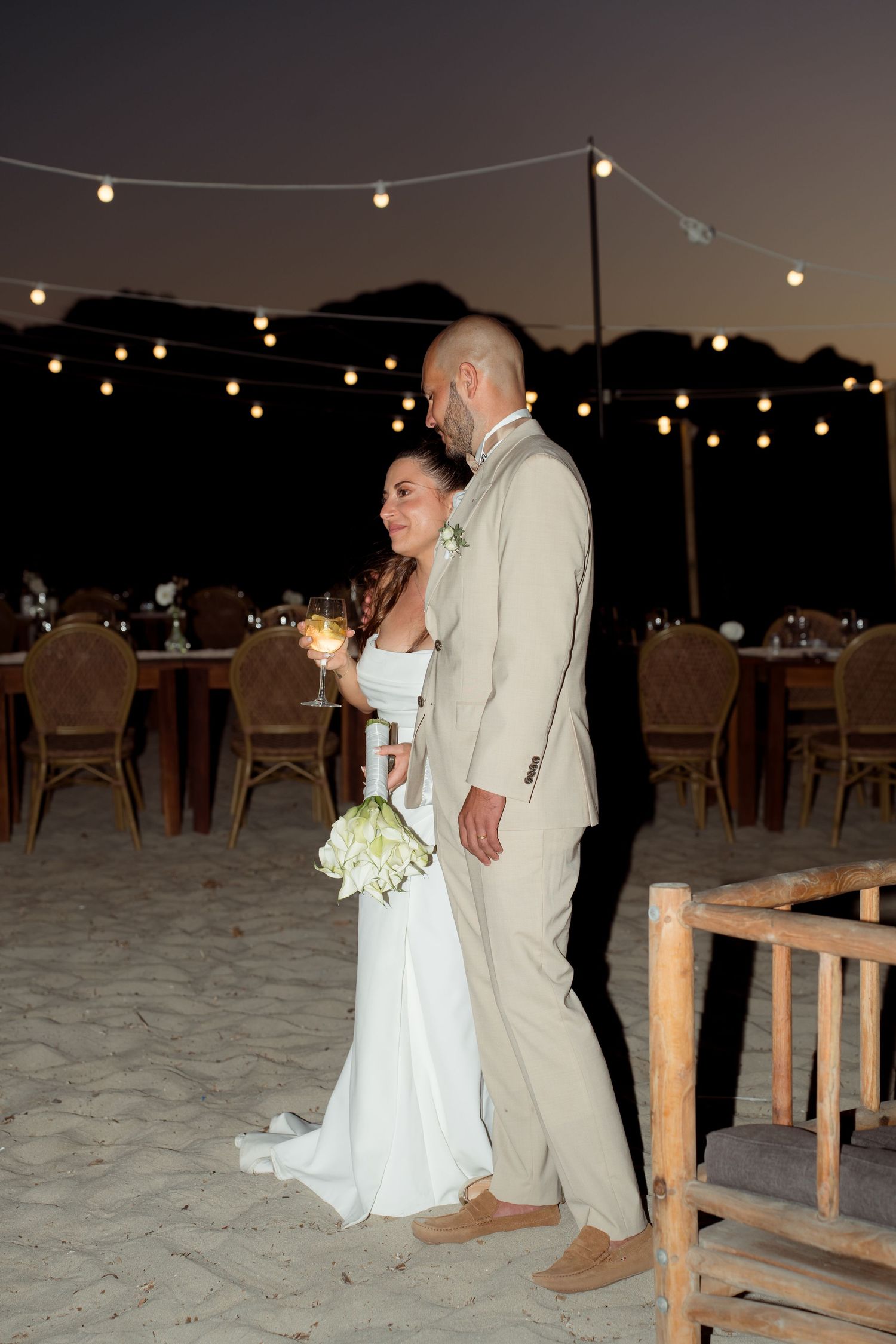 Newlyweds embrace on sandy beach at night with glowing string lights and reception tables behind them.