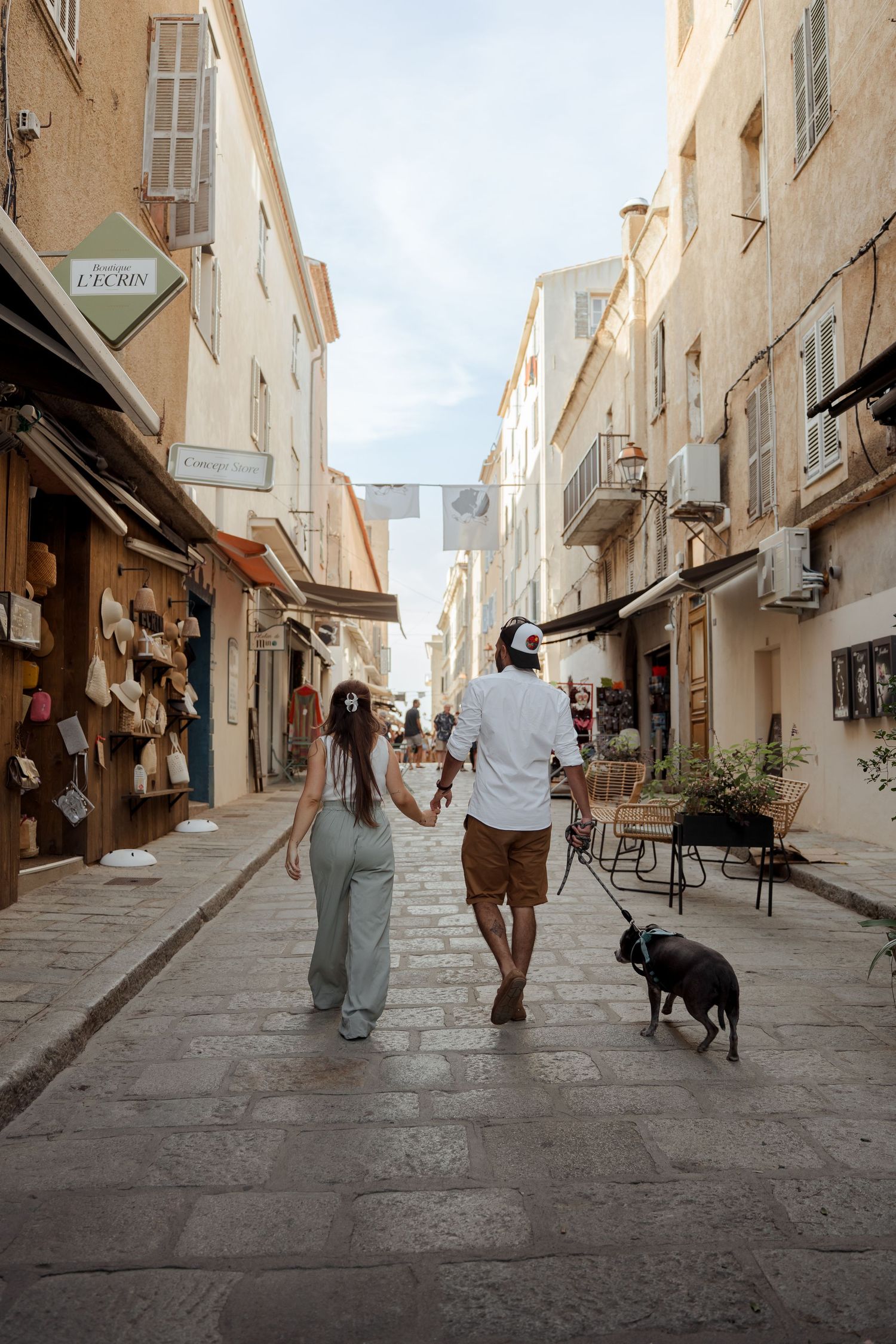 Couple walking hand in hand with a black dog through a charming European cobblestone street lined with shops.