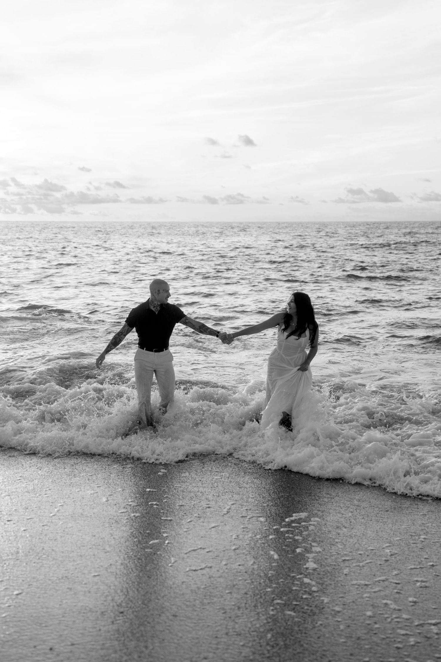 Black and white photo of couple holding hands in crashing ocean waves on a beach at sunset.
