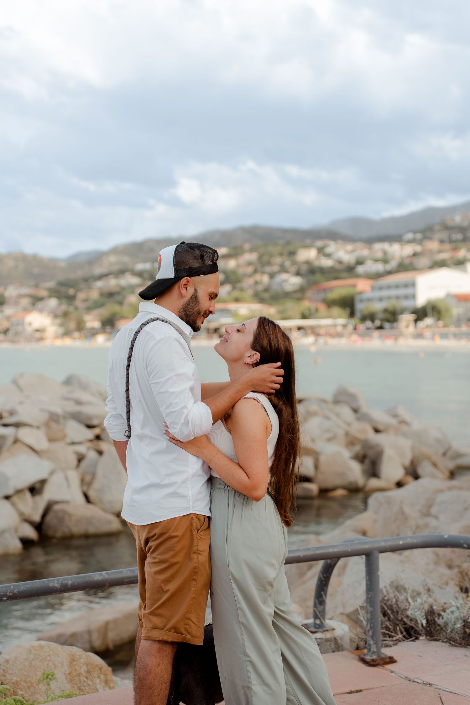 Loving couple about to kiss near rocky waterfront with a scenic coastal town and mountains behind them.