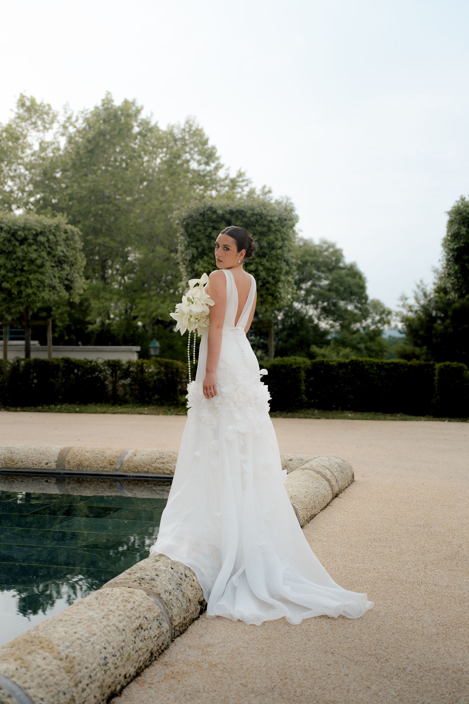 Bride in backless white gown holding floral bouquet beside garden pool.