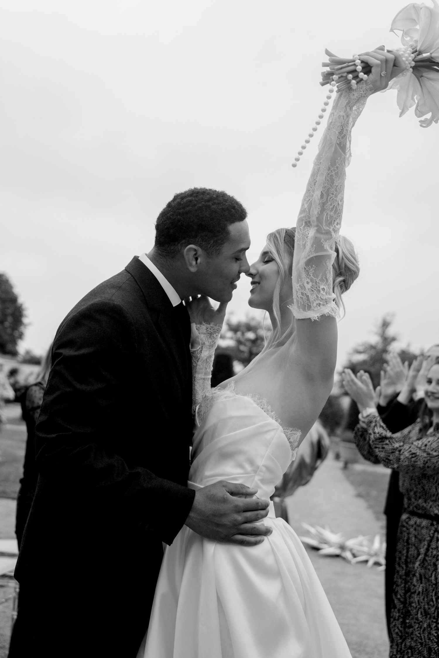Black and white close-up of bride and groom kissing, bride raising bouquet and veil triumphantly after wedding ceremony.