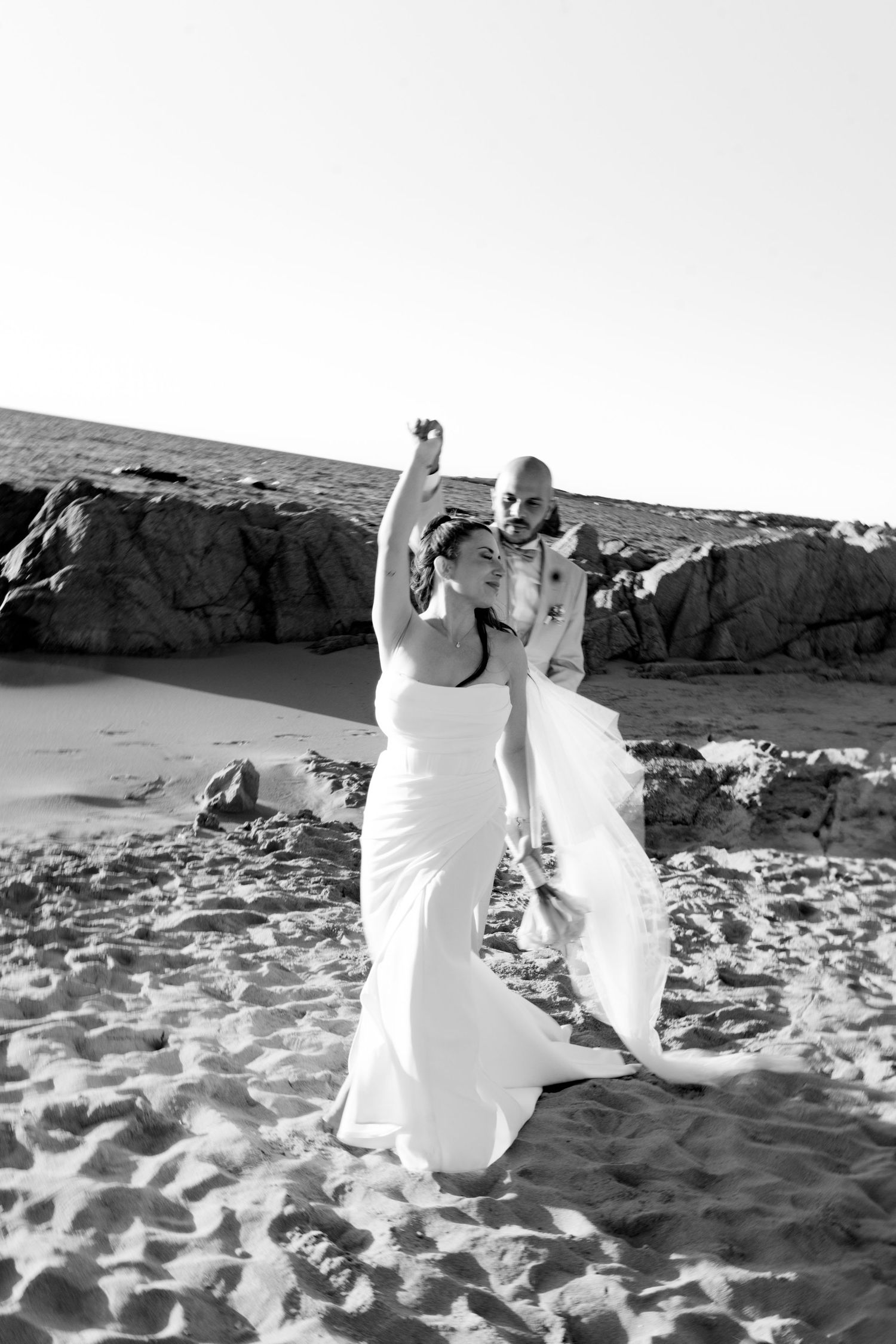 Black and white photo of bride and groom celebrating with raised arms on sandy beach at sunset.